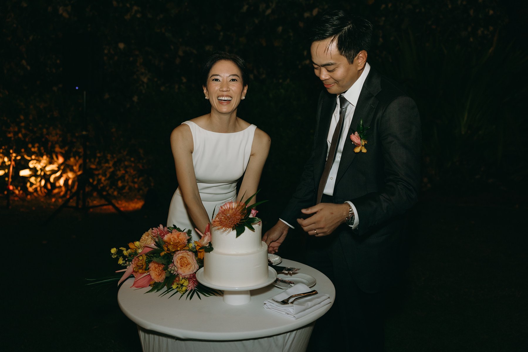 Couple cutting wedding cake with tropical flowers during evening reception at resort wedding venue