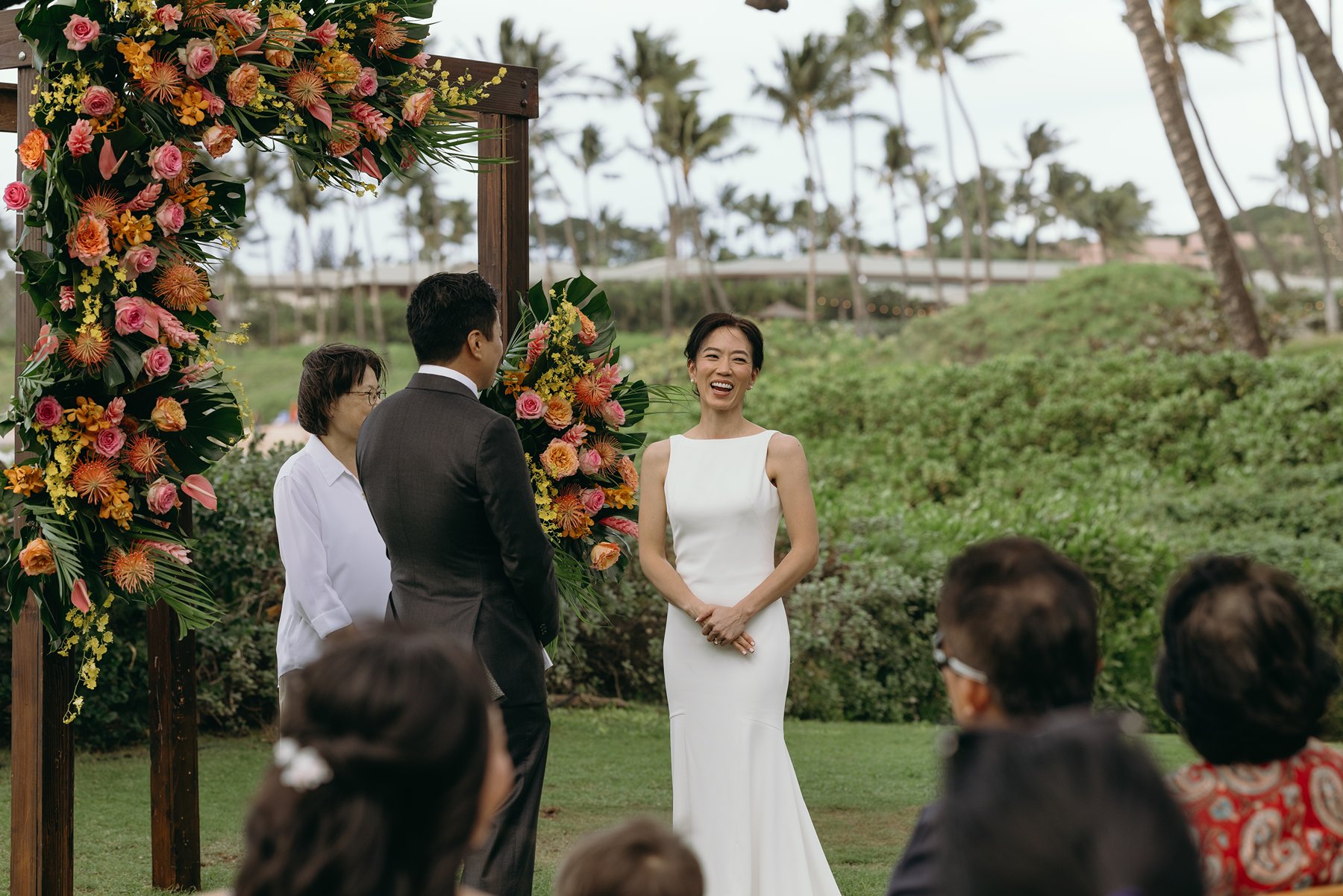 Bride smiling during wedding ceremony under tropical floral arch with ocean view