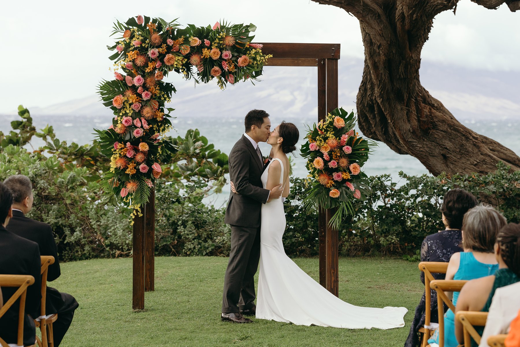 Bride and groom kissing under floral arch during oceanfront ceremony at resort wedding venue in Hawaii