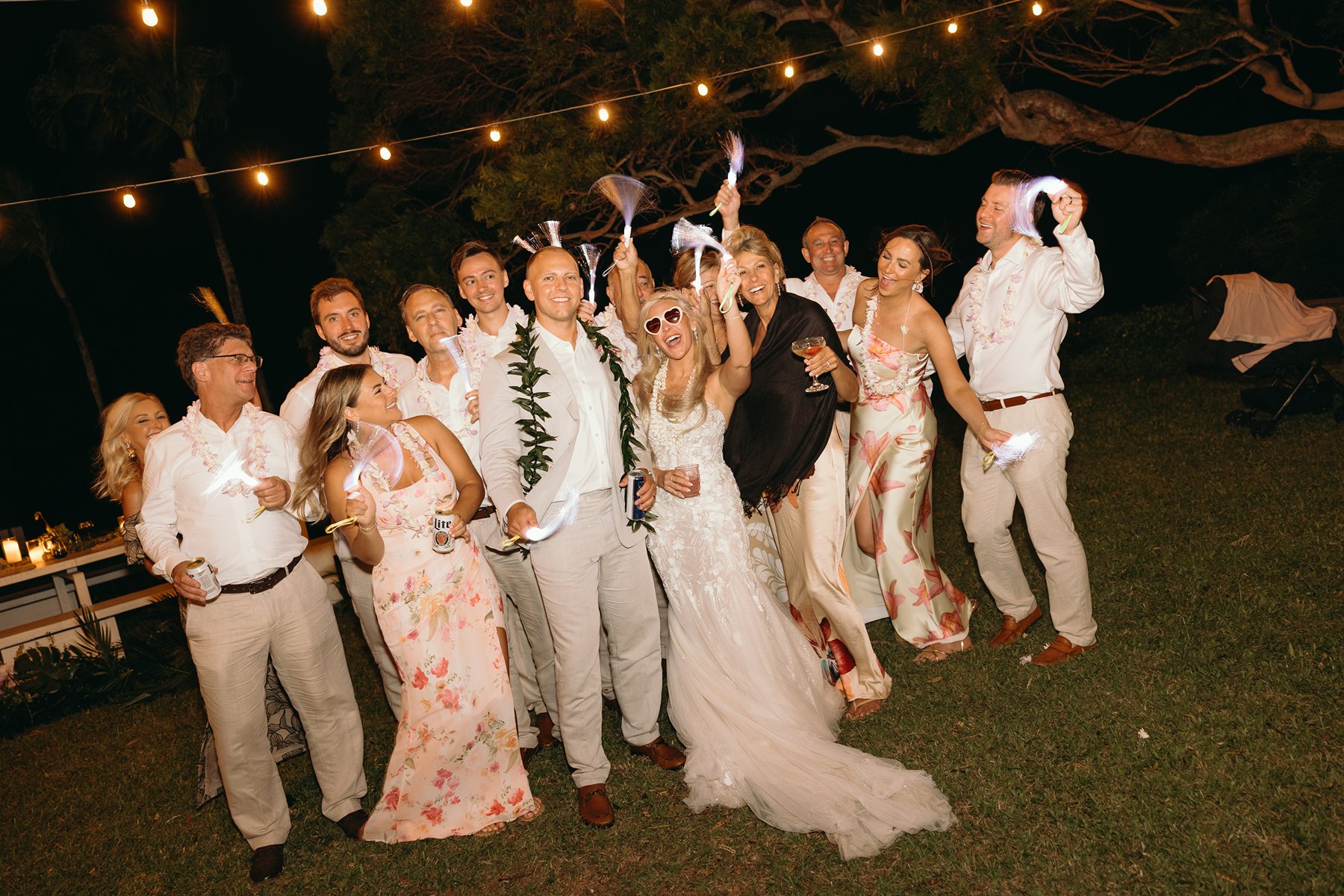 Joyful wedding reception under string lights as the bride and groom dance with friends on the lawn, guests cheering and waving napkins in the warm Maui night air.
