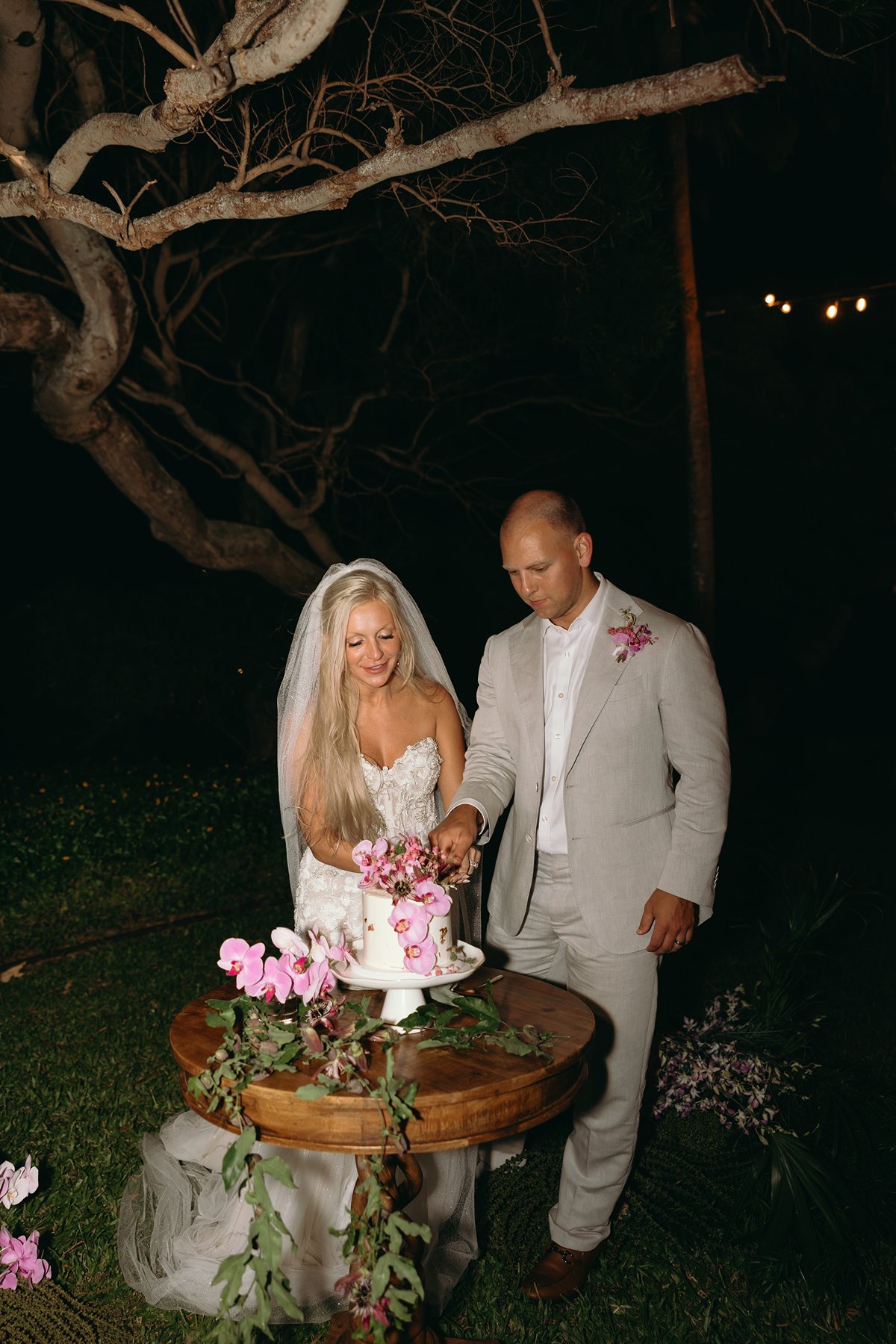Bride and groom cutting their orchid-covered cake after they elope in Maui, standing beneath twisted tree branches on a candlelit lawn.