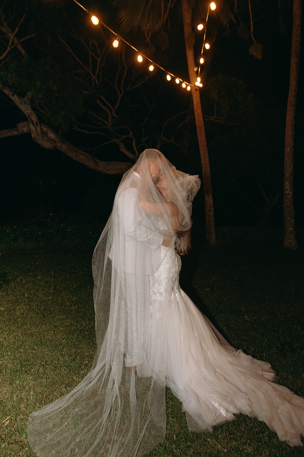 Bride and groom sharing a romantic kiss beneath string lights after they elope in Maui, her veil floating softly in the night air.
