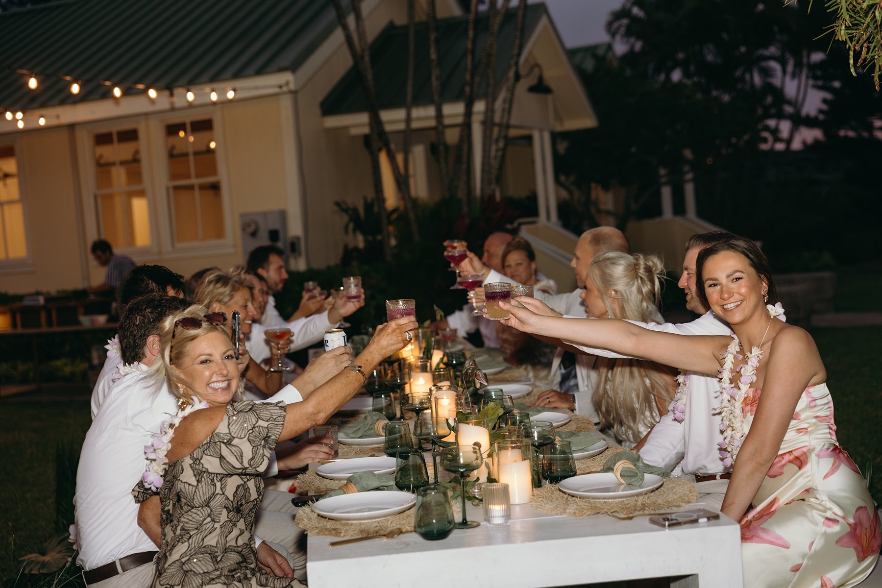 Guests raising cocktails in a candlelit toast during a warm Maui evening reception, string lights glowing overhead.