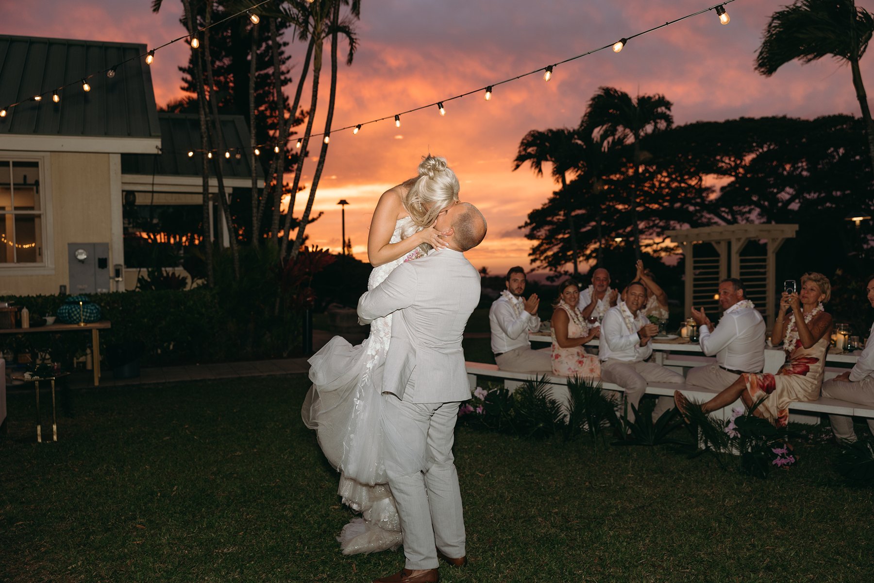 Groom lifting his bride at sunset during their elope in Maui, golden sky glowing behind palm trees while guests applaud from nearby tables.