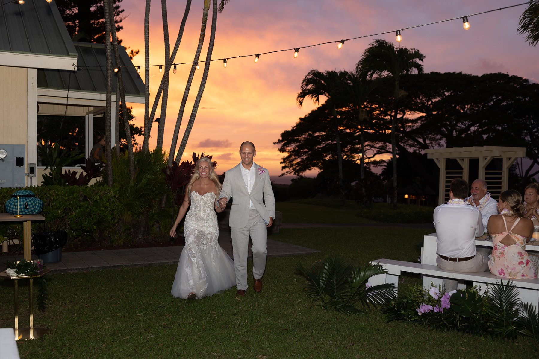 Newlyweds walking hand in hand across a Maui resort lawn at sunset after they chose to elope in Maui, string lights glowing overhead.