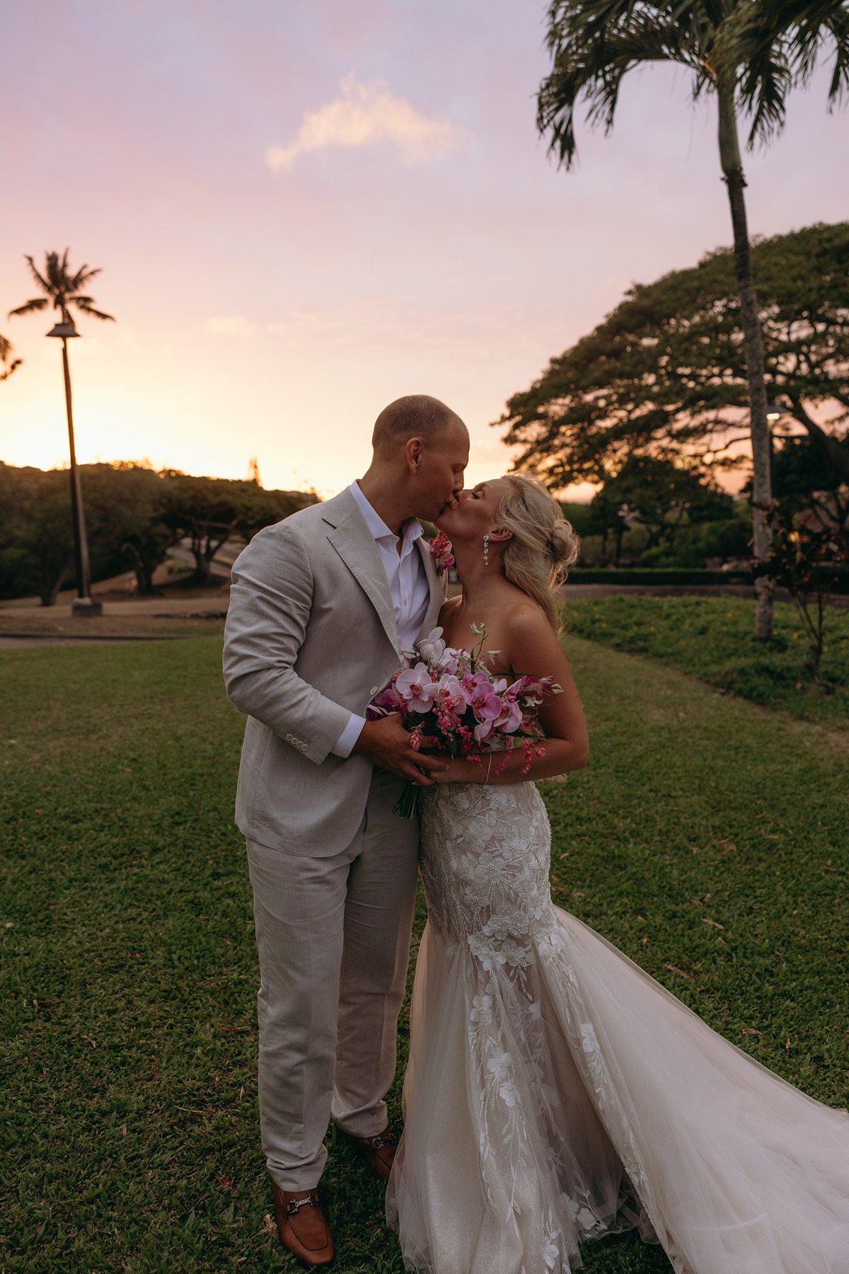 Bride and groom sharing a quiet kiss on a grassy lawn at golden hour, her pink bouquet glowing softly in the Maui sunset.