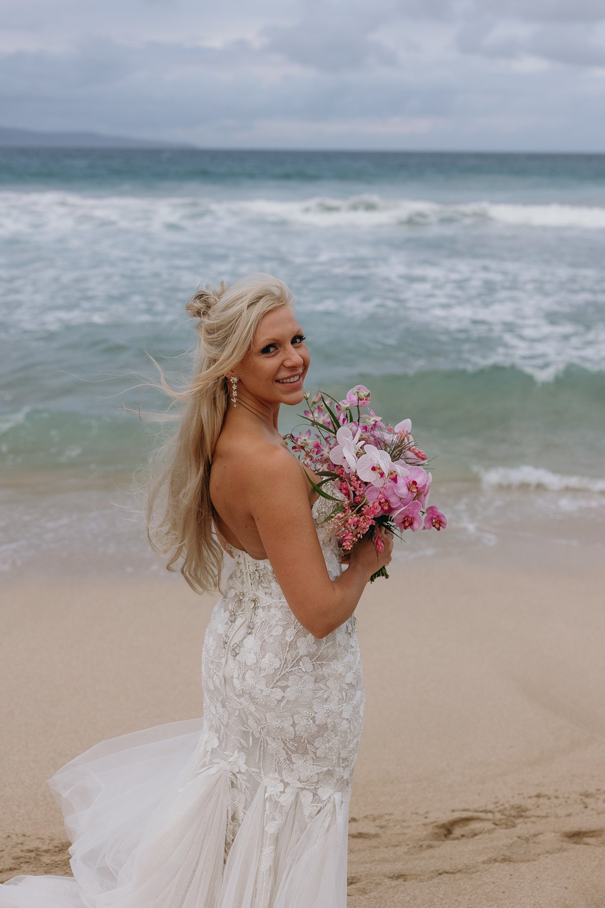 Bride holding a pink tropical bouquet at the water’s edge after choosing to elope in Maui, wind catching her hair as waves crash behind her.