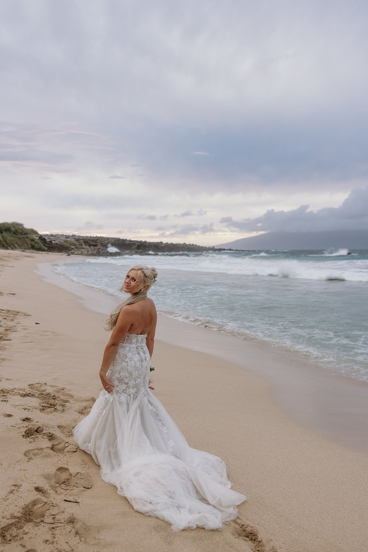 Bride walking barefoot along a quiet shoreline, dress trailing in the sand as she celebrates her decision to elope in Maui.