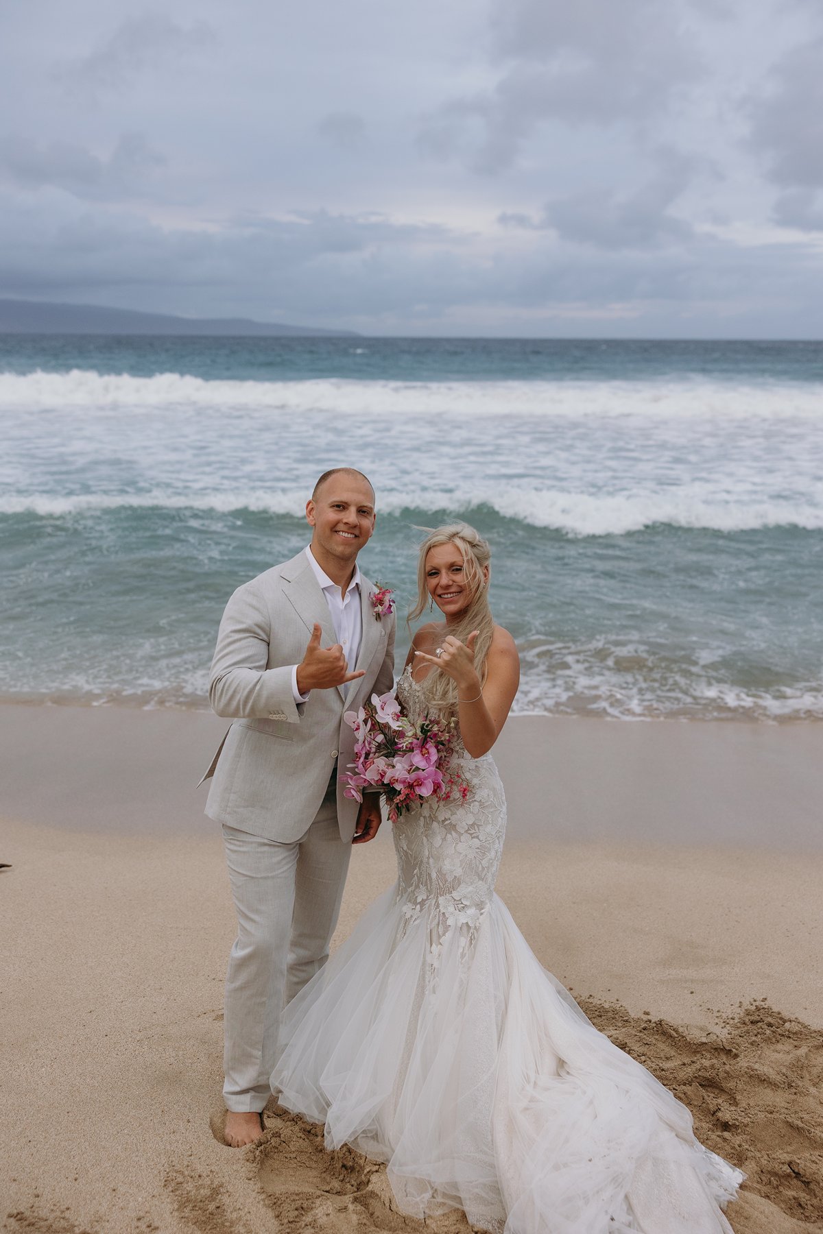 Smiling bride and groom flashing shaka signs at the shoreline, bouquet bright against the soft blue ocean.