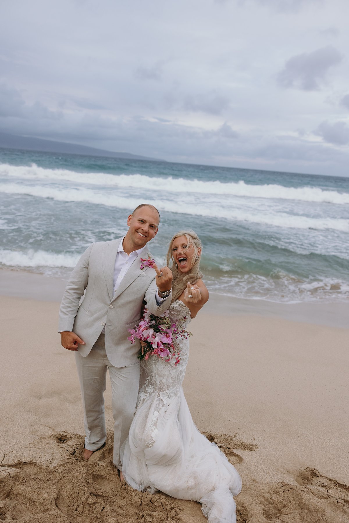 Bride and groom celebrating barefoot on a windy beach after choosing to elope in Maui, waves crashing behind them as she holds a bright pink tropical bouquet.