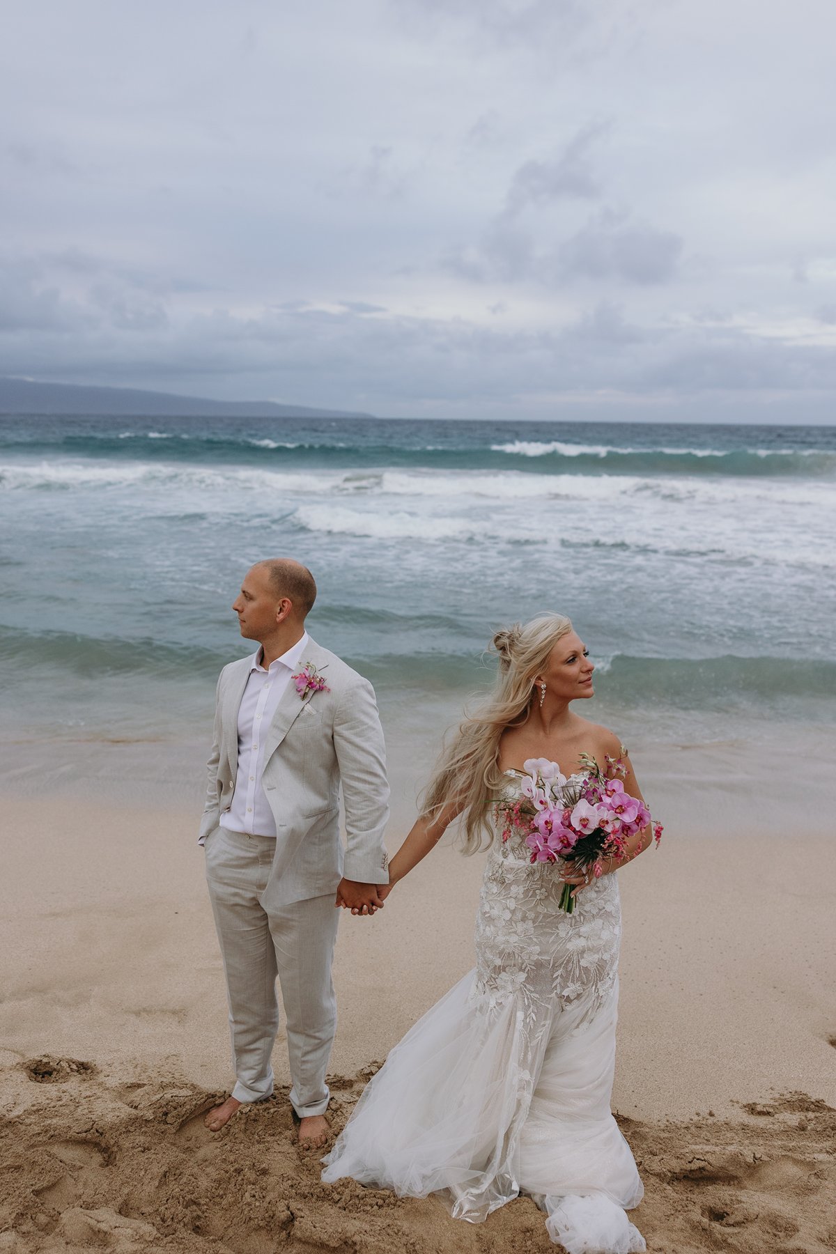 Bride and groom standing hand in hand on the beach after they elope in Maui, looking opposite directions as the ocean swells behind them.