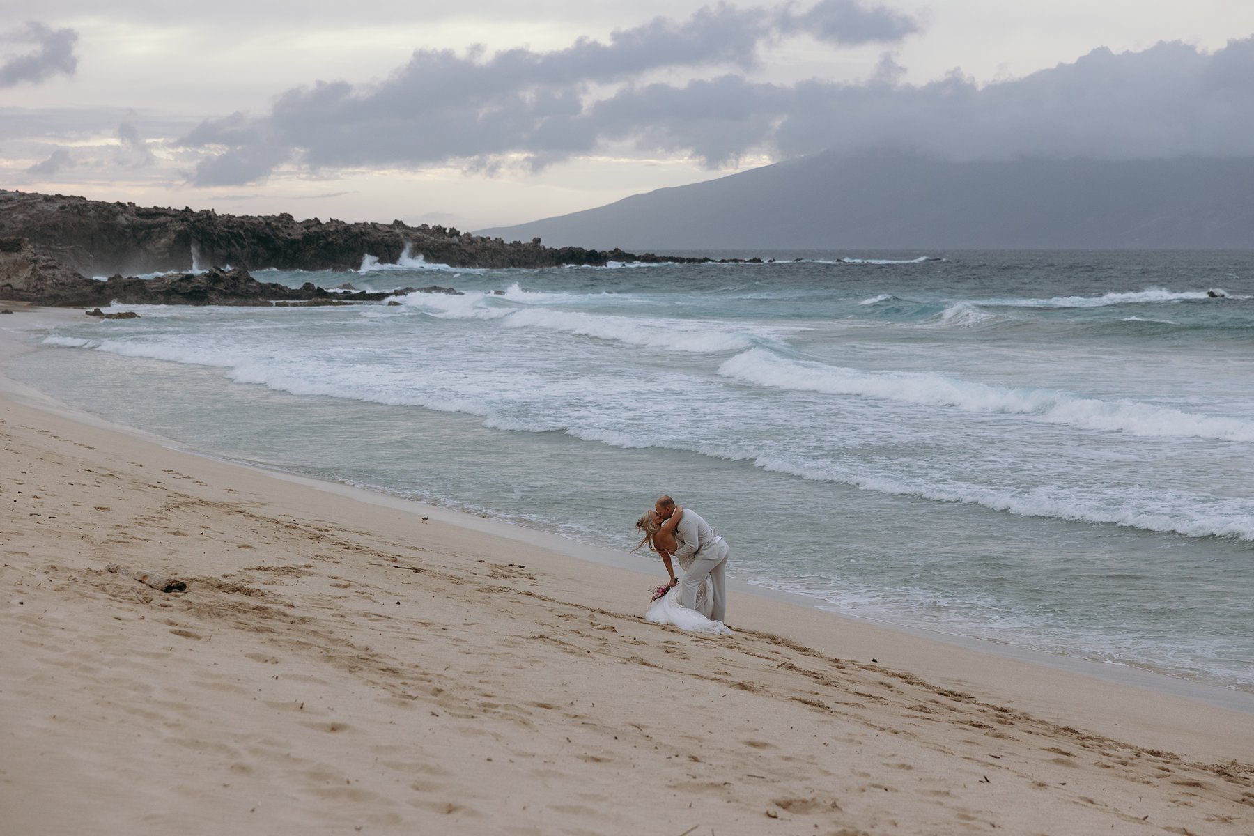 Newlyweds embracing on a windswept beach after they elope in Maui, waves rolling in under a moody island sky.