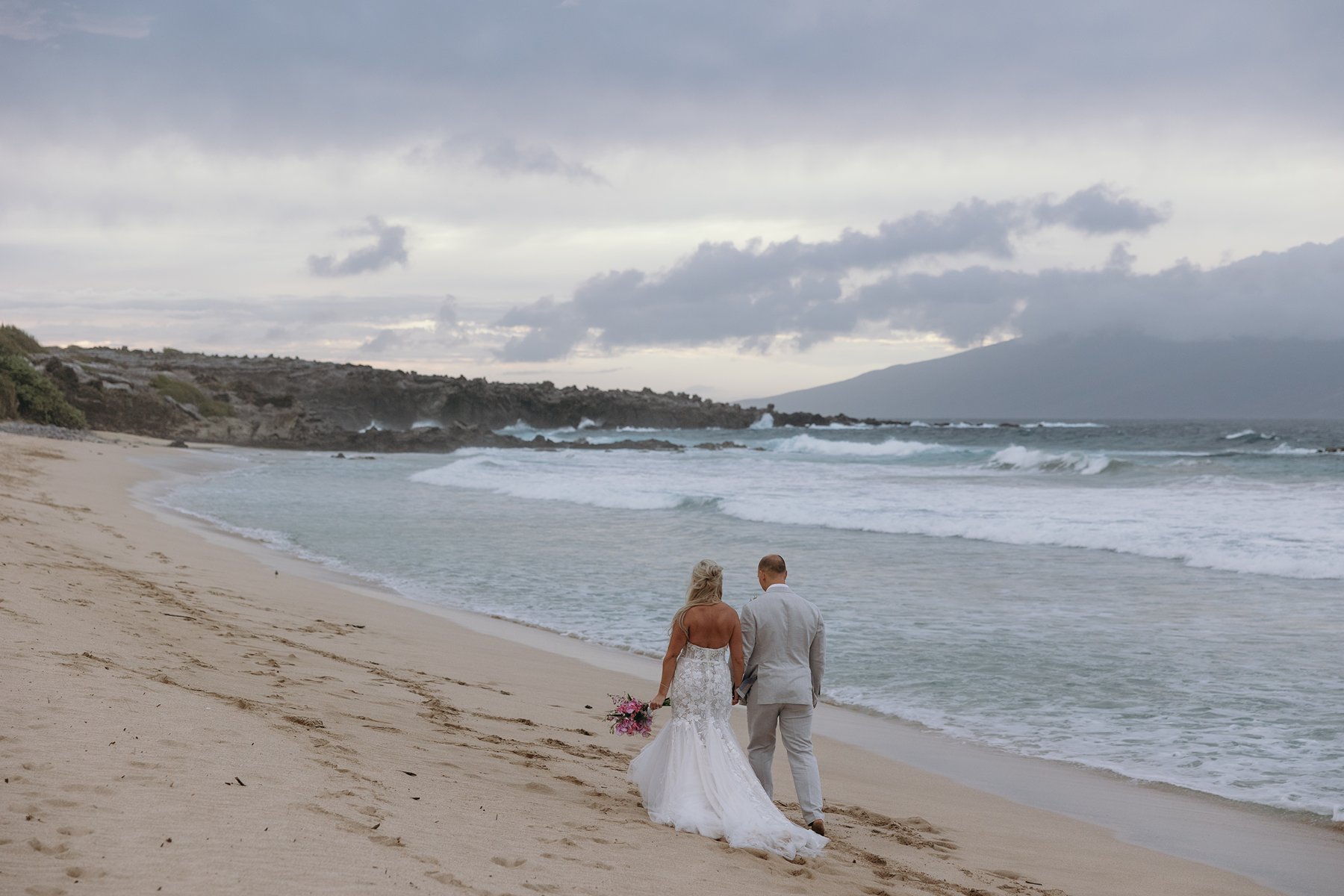 Newlyweds strolling barefoot along the shoreline after choosing to elope in Maui, waves rolling in under a soft, cloudy sky.