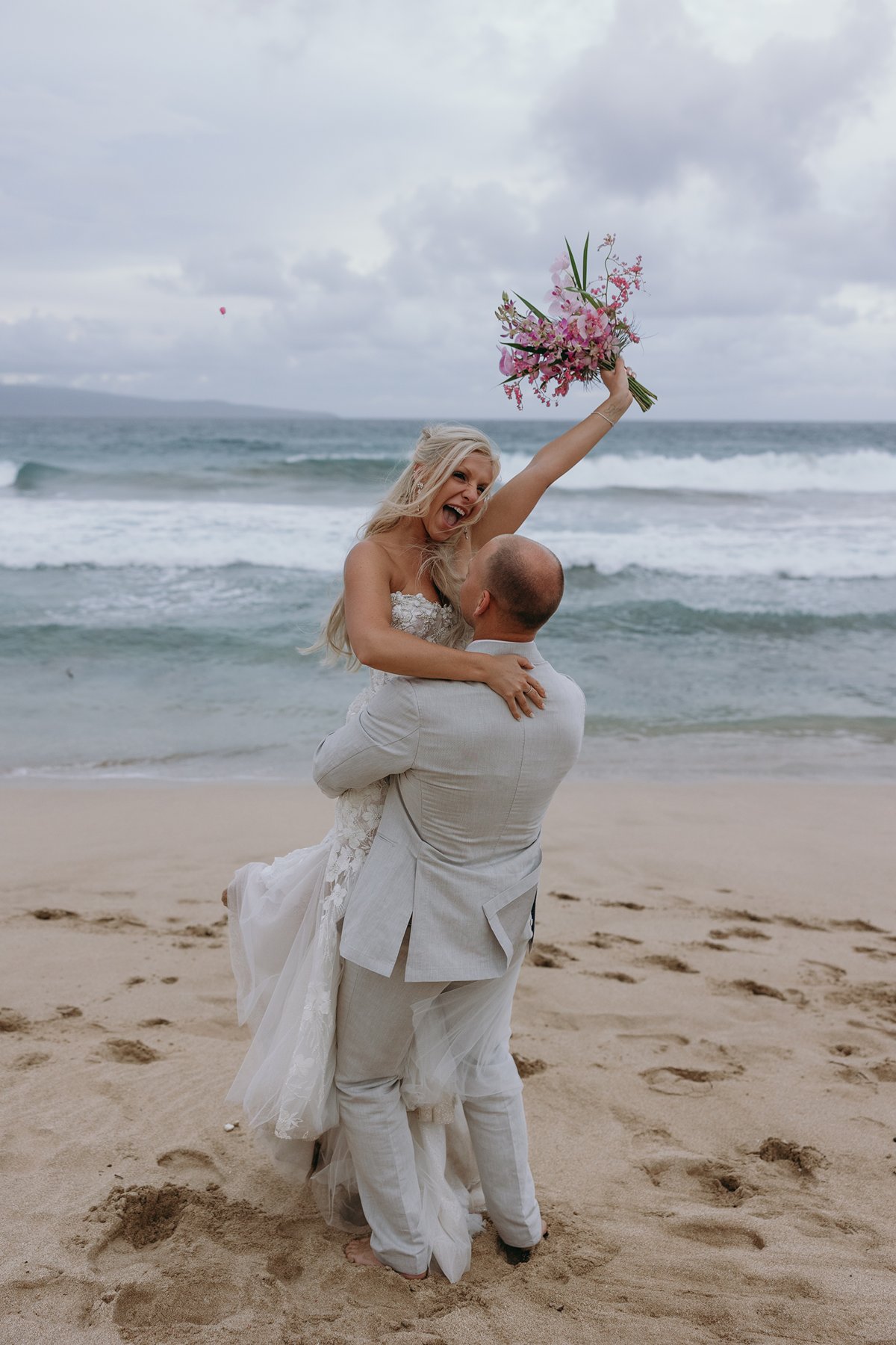 Groom lifting his bride in celebration as they elope in Maui, her bouquet raised high with white-capped waves in the background.