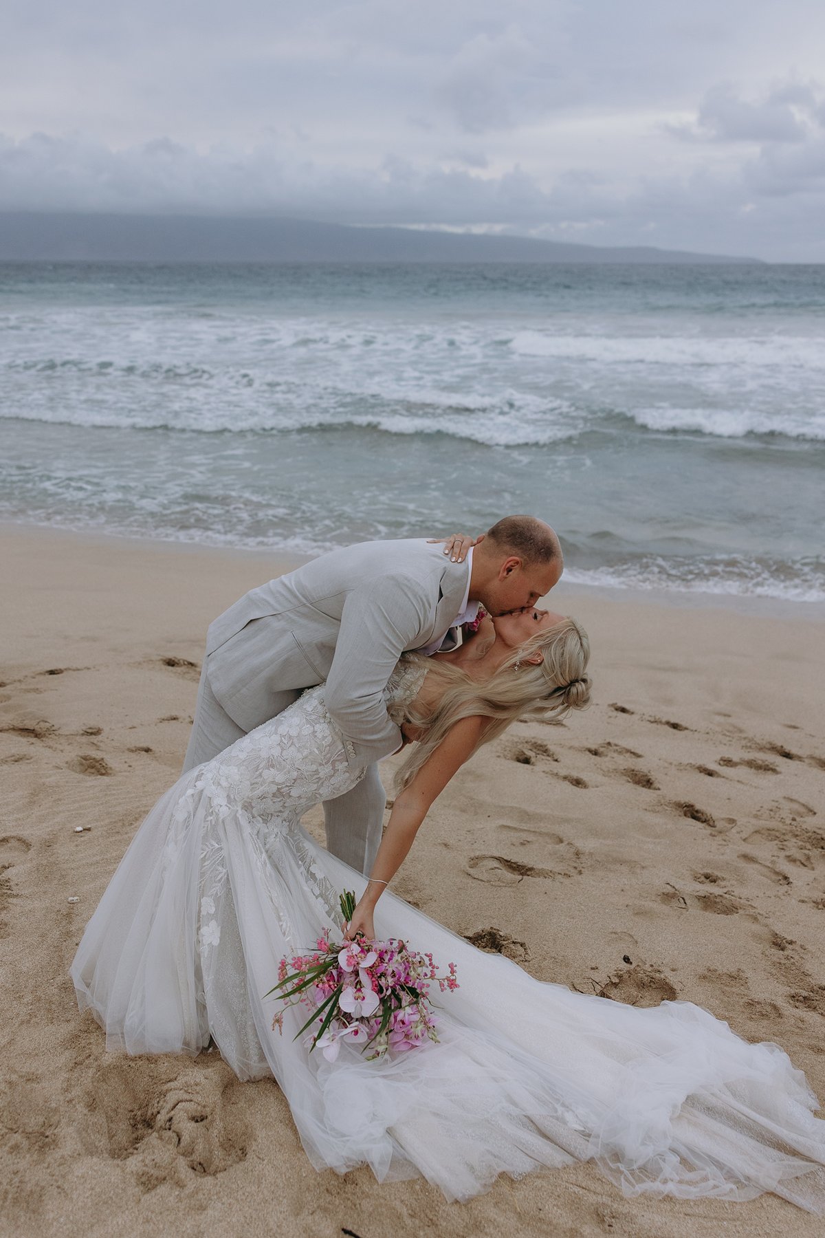 Bride and groom sharing a sunset dip and kiss on the beach after choosing to elope in Maui, waves rolling onto the sand behind them.