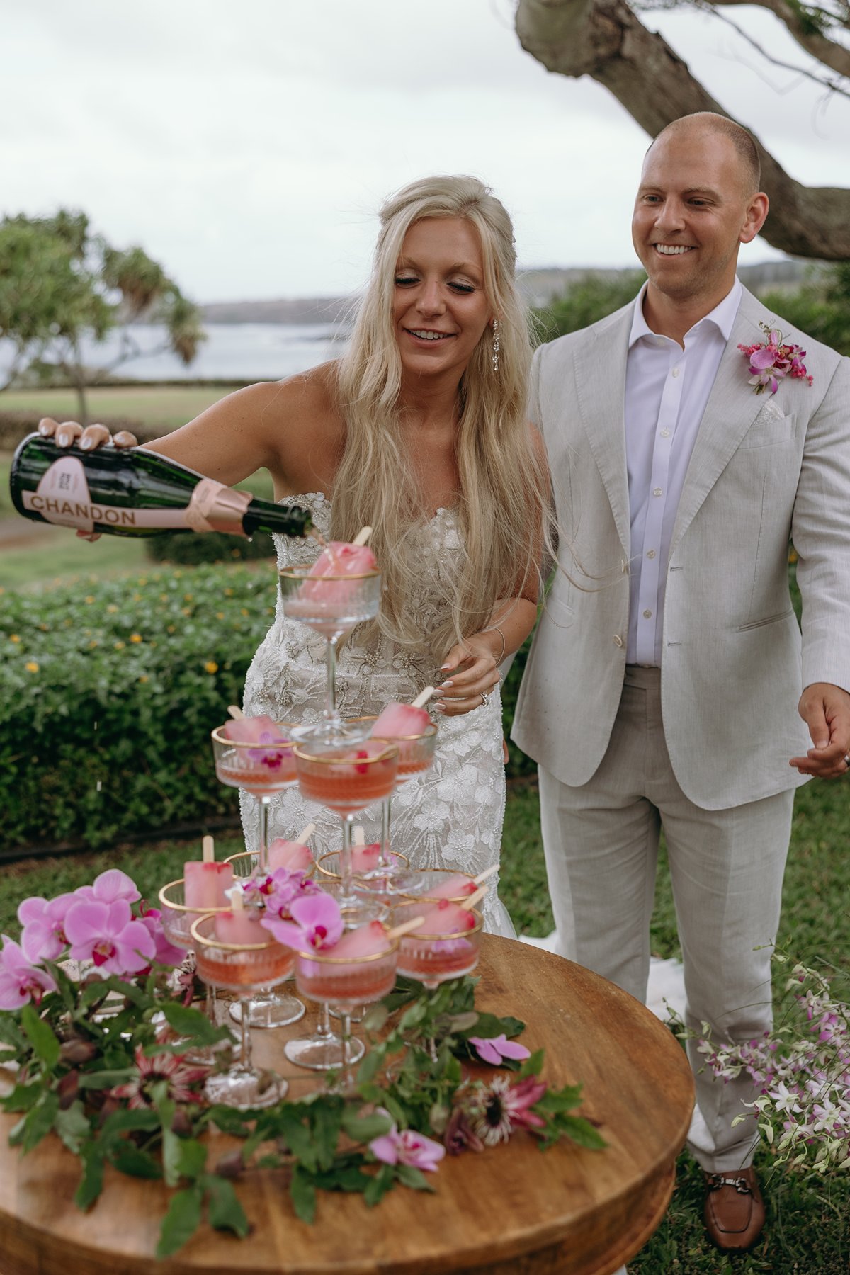 Bride pouring champagne into coupe glasses topped with pink florals during an intimate celebration after they elope in Maui.