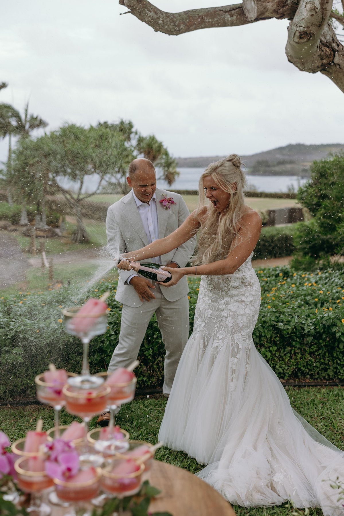 Bride popping champagne into a tower of pink cocktails after choosing to elope in Maui, ocean and greenery stretching behind them.