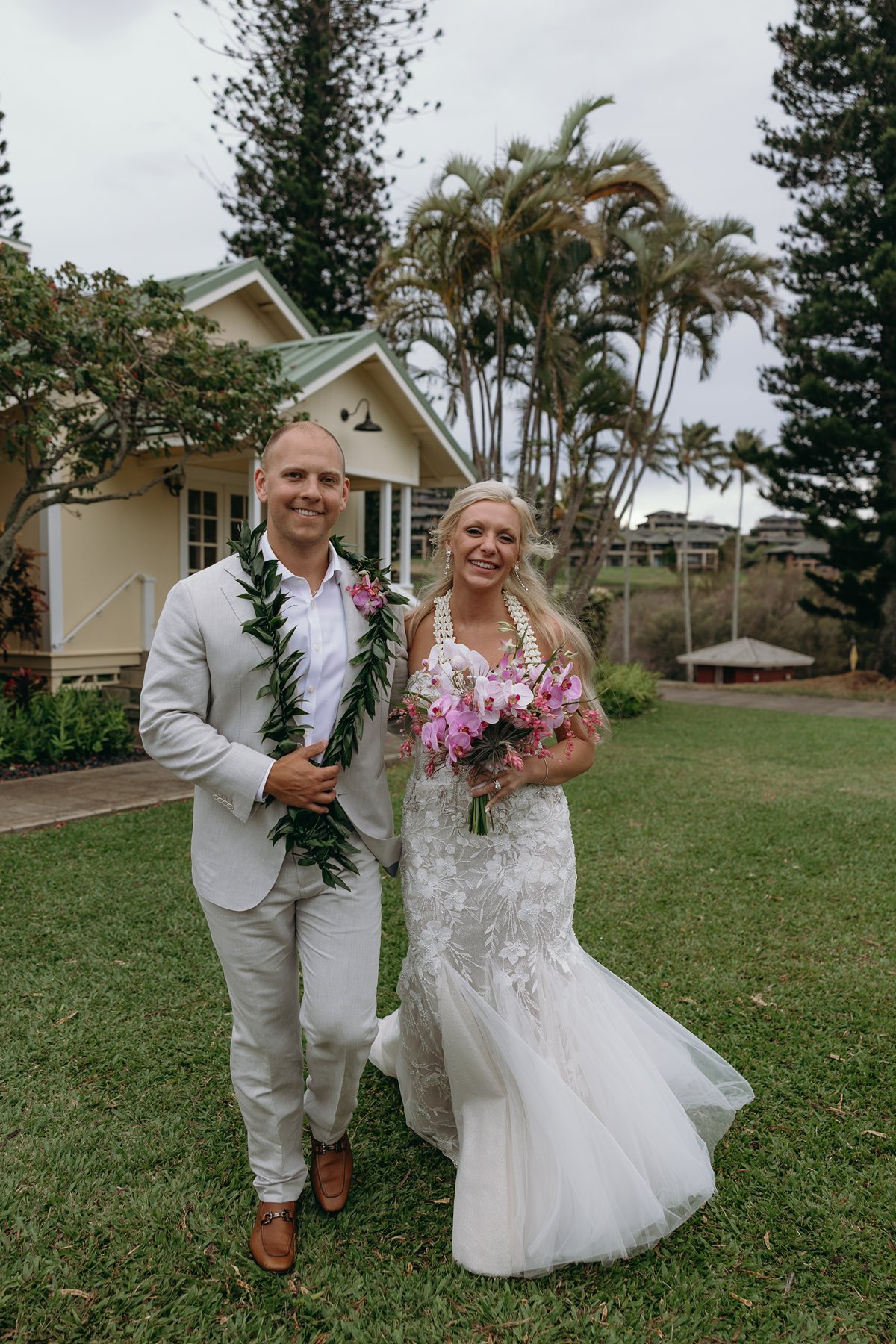 Bride and groom walking across a green lawn after they elope in Maui, her lace dress flowing and his maile lei resting over a light gray suit.