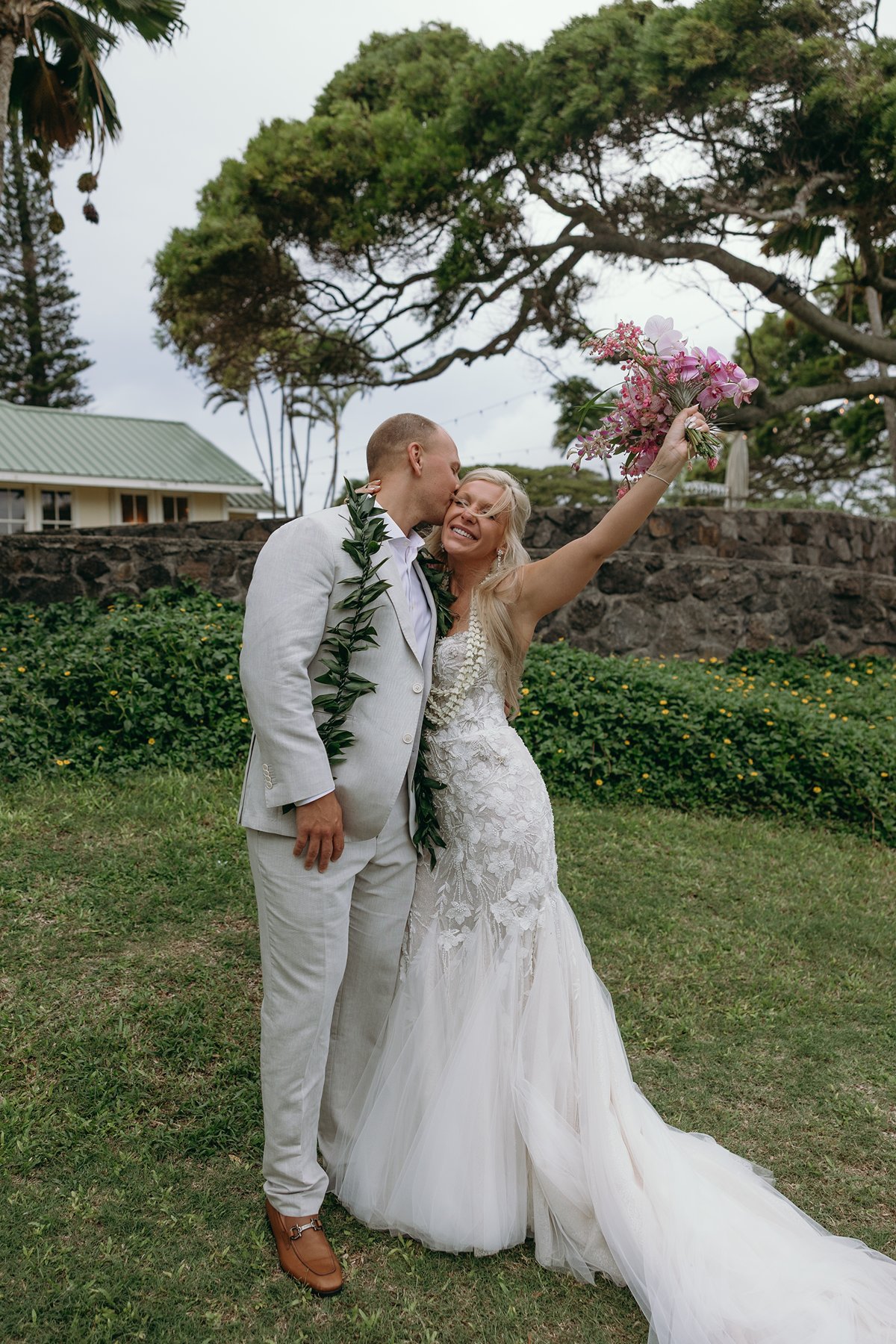 Groom kissing his bride on the cheek as she lifts her bouquet in celebration after they elope in Maui, tropical greenery surrounding them.