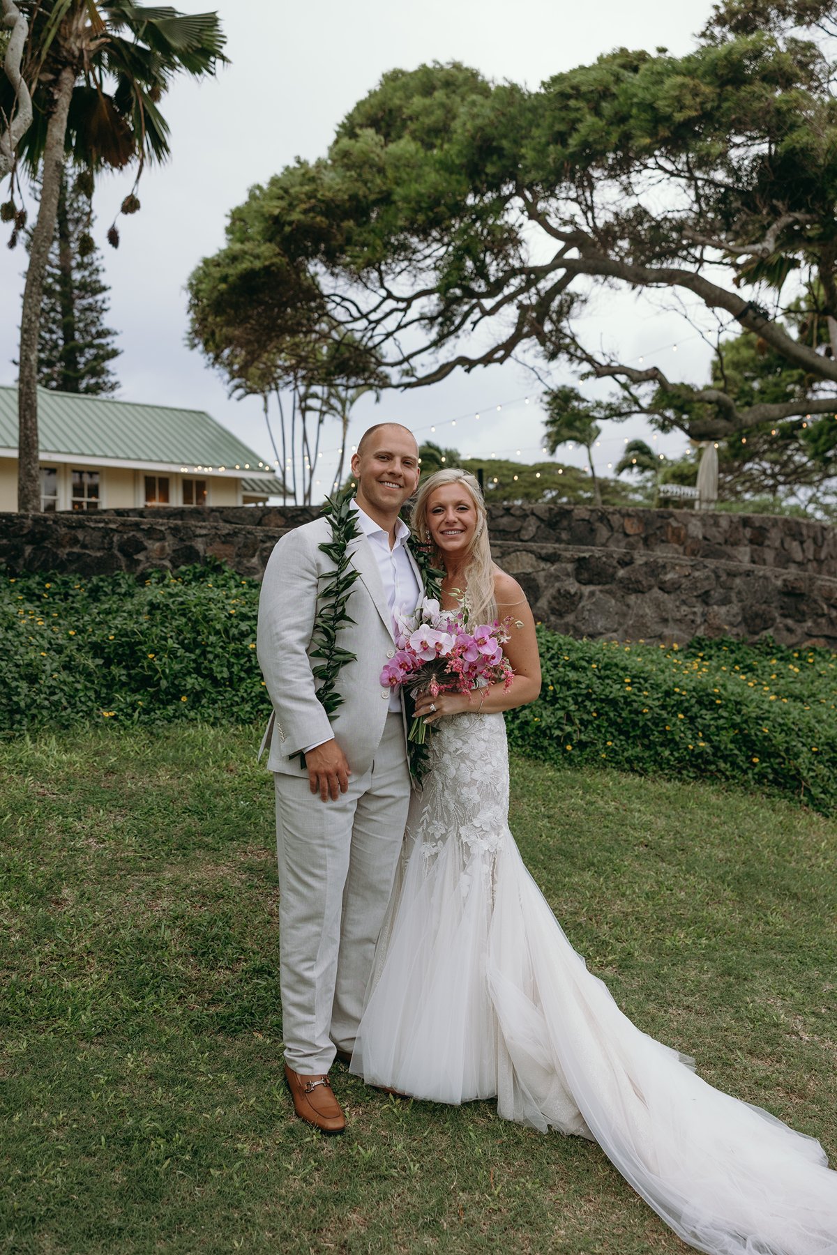 Newlyweds smiling at the camera on a green lawn after they elope in Maui, pink bouquet and maile lei adding tropical color.
