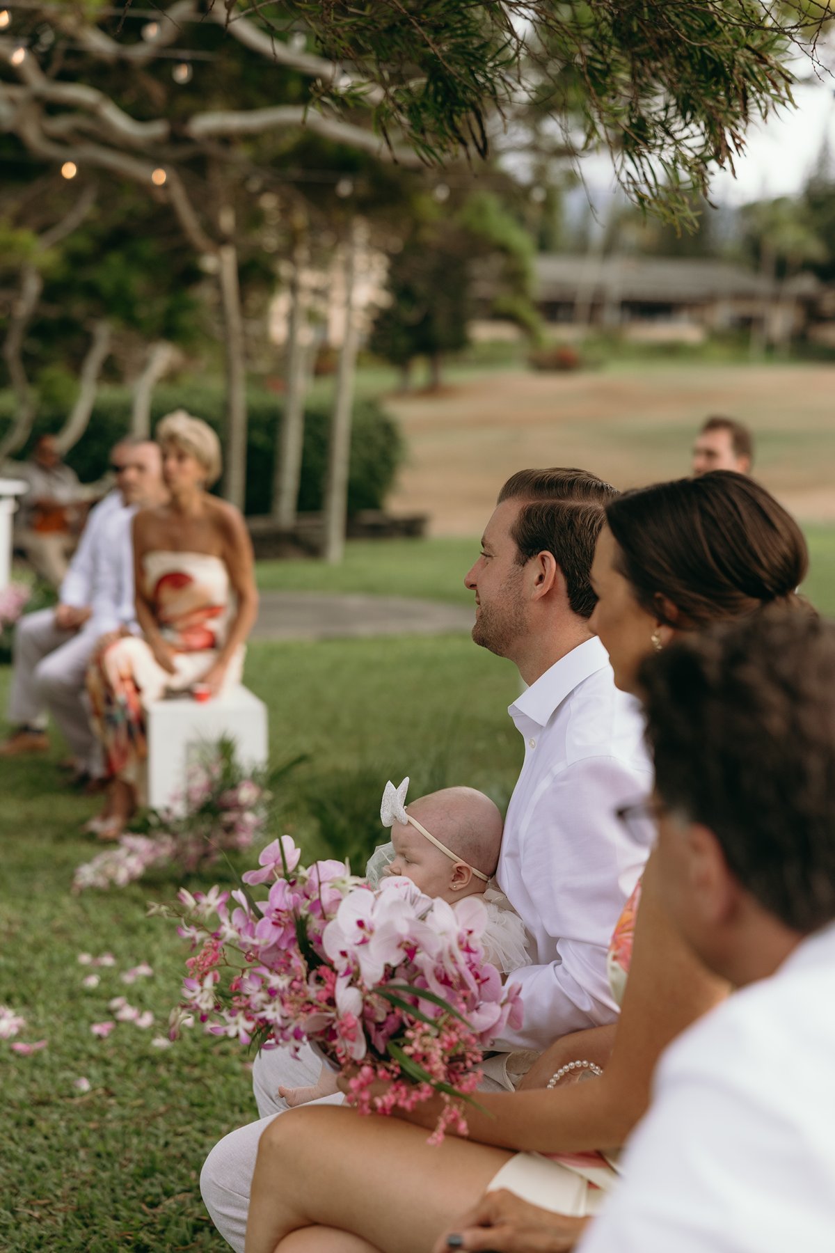 Family members smiling during an outdoor Maui ceremony, baby holding a bouquet of pink orchids in the front row.