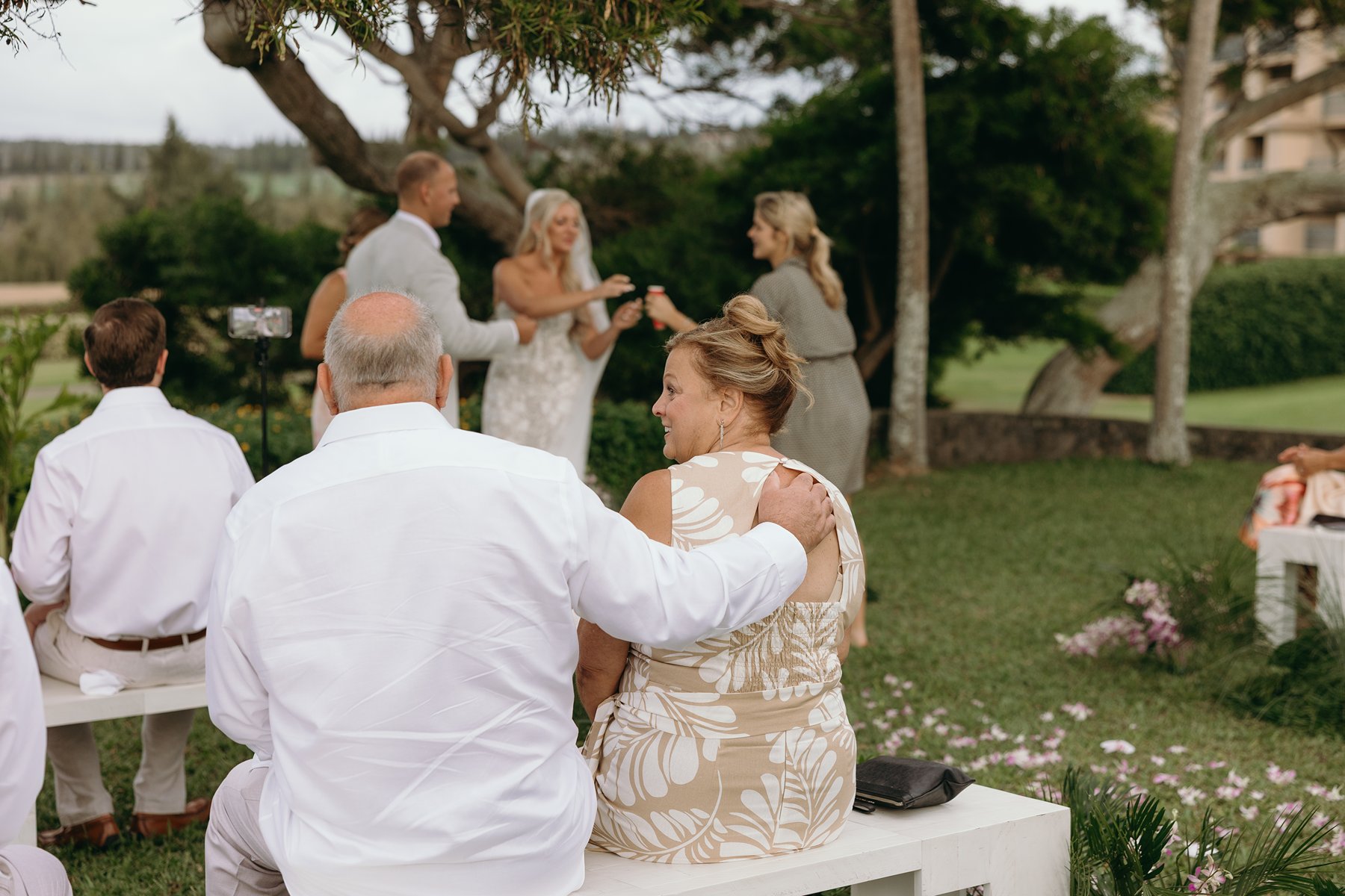 Guests seated on white benches watching the ceremony unfold, pink petals scattered across the grass.
