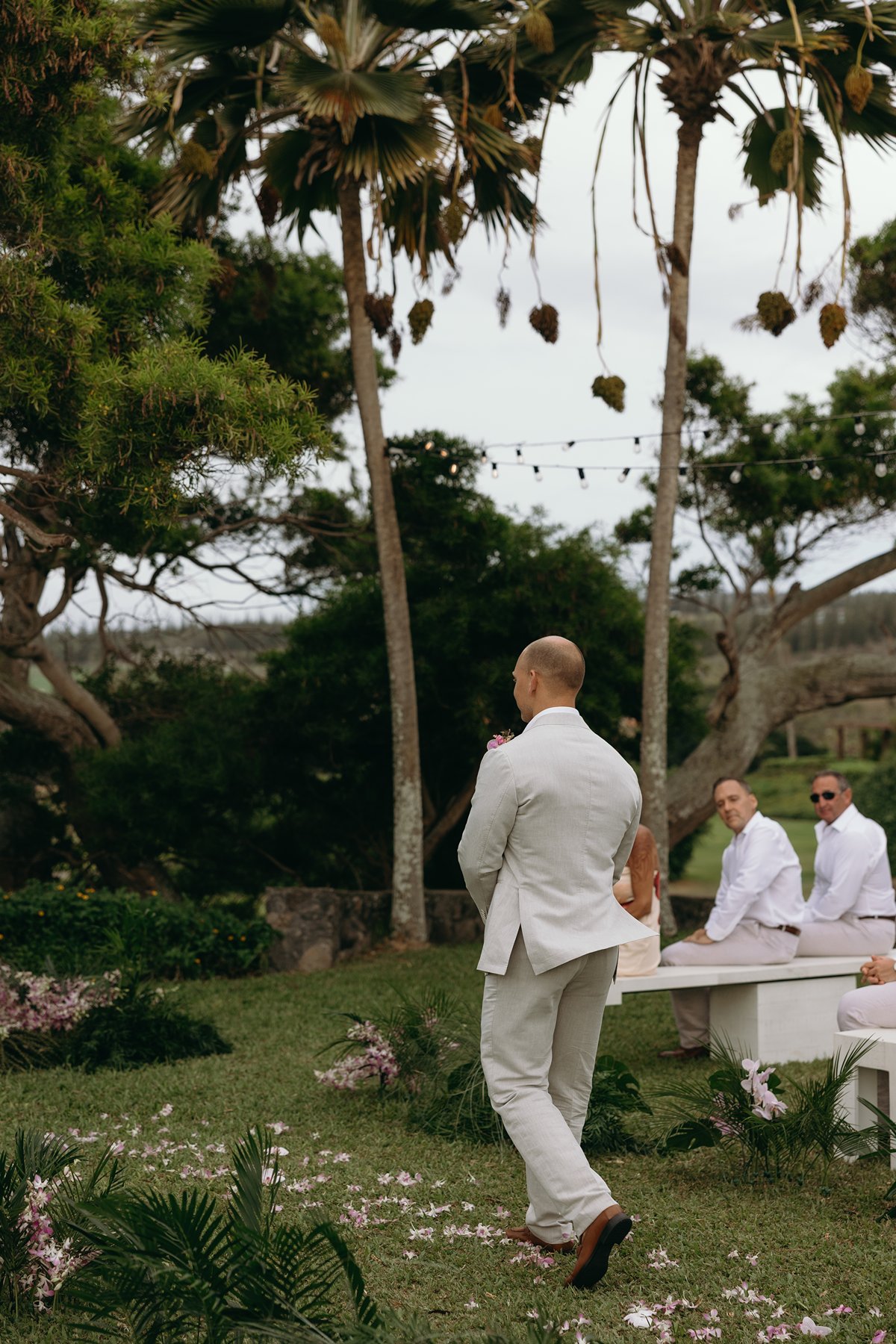 Groom walking across a garden lawn toward the ceremony site as guests wait beneath swaying palm trees and string lights.
