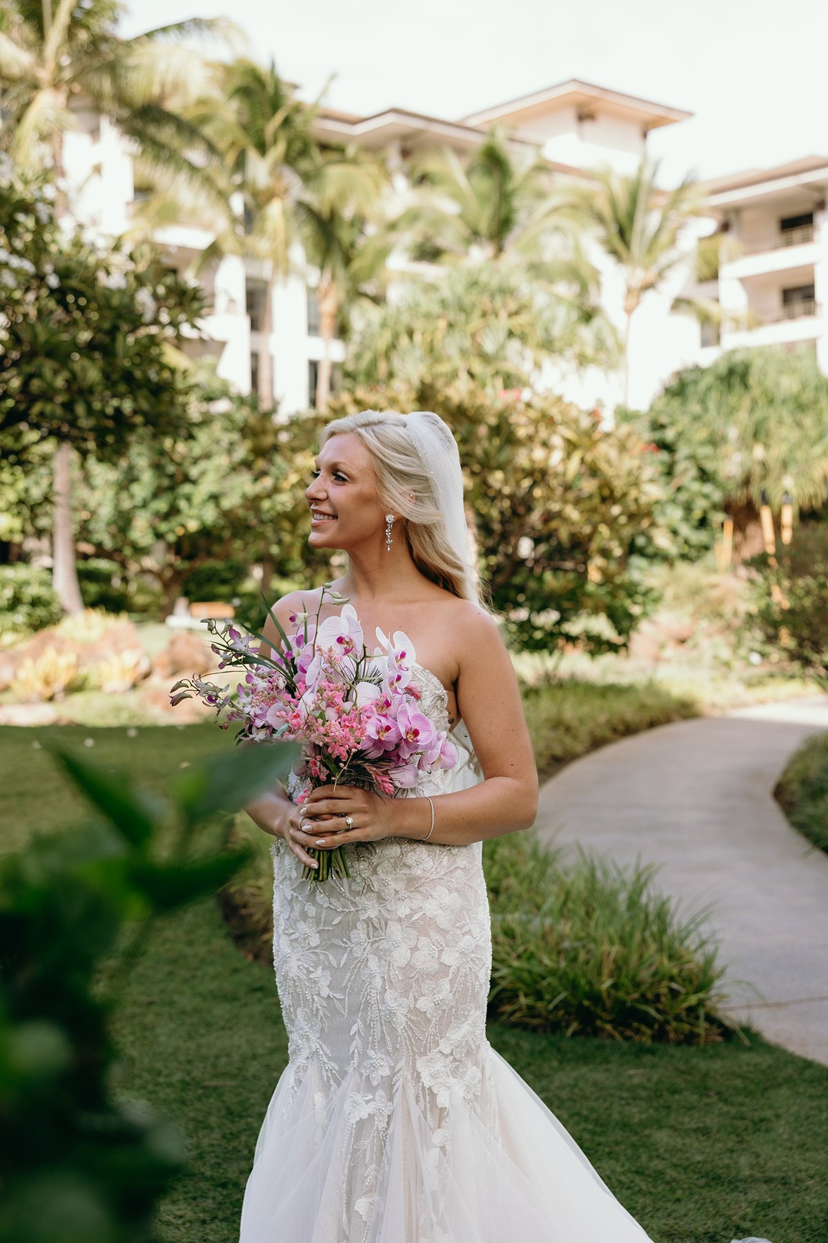 Bride holding a vibrant pink bouquet in a lush resort garden after she chose to elope in Maui, palm trees swaying behind her.