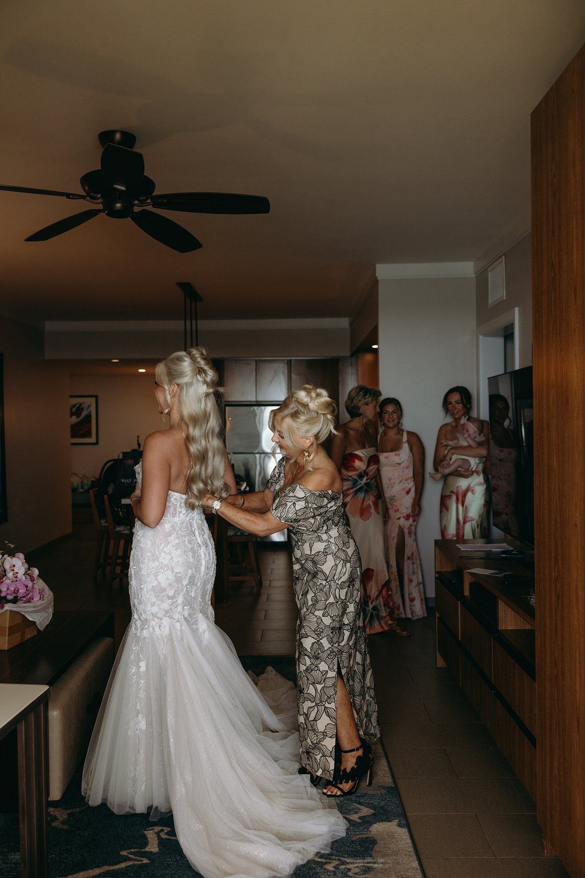 Mother fastening the back of the bride’s dress inside a softly lit room before the couple elope in Maui, bridesmaids watching nearby.