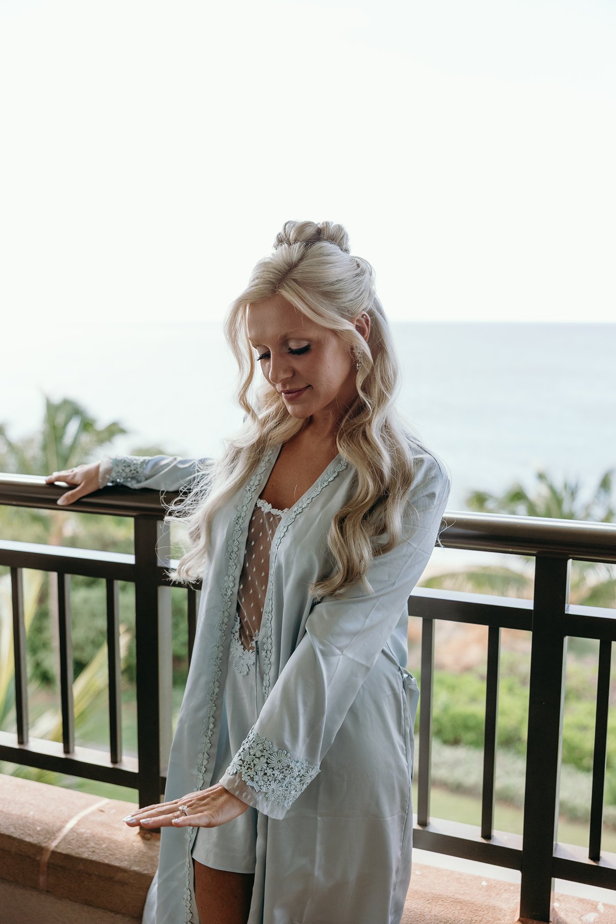 Bride leaning against a balcony railing in a soft blue robe, ocean and palm trees stretching out behind her.