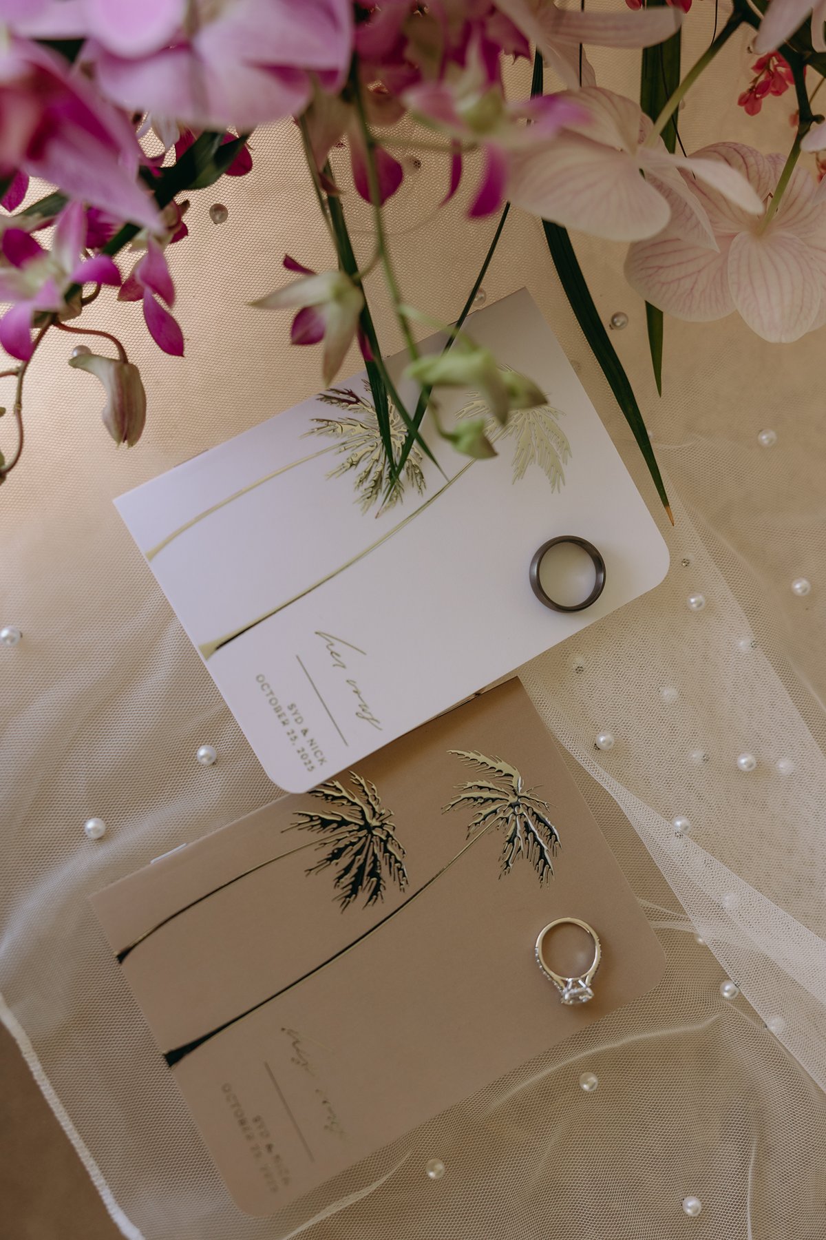 Flat lay of wedding invitations and rings surrounded by orchids and soft tulle, styled for a romantic Maui celebration.