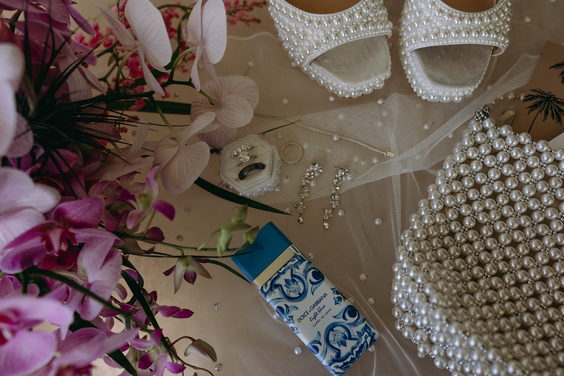 Bridal details featuring pearl heels, jewelry, perfume, and a beaded clutch arranged beside pink tropical florals.