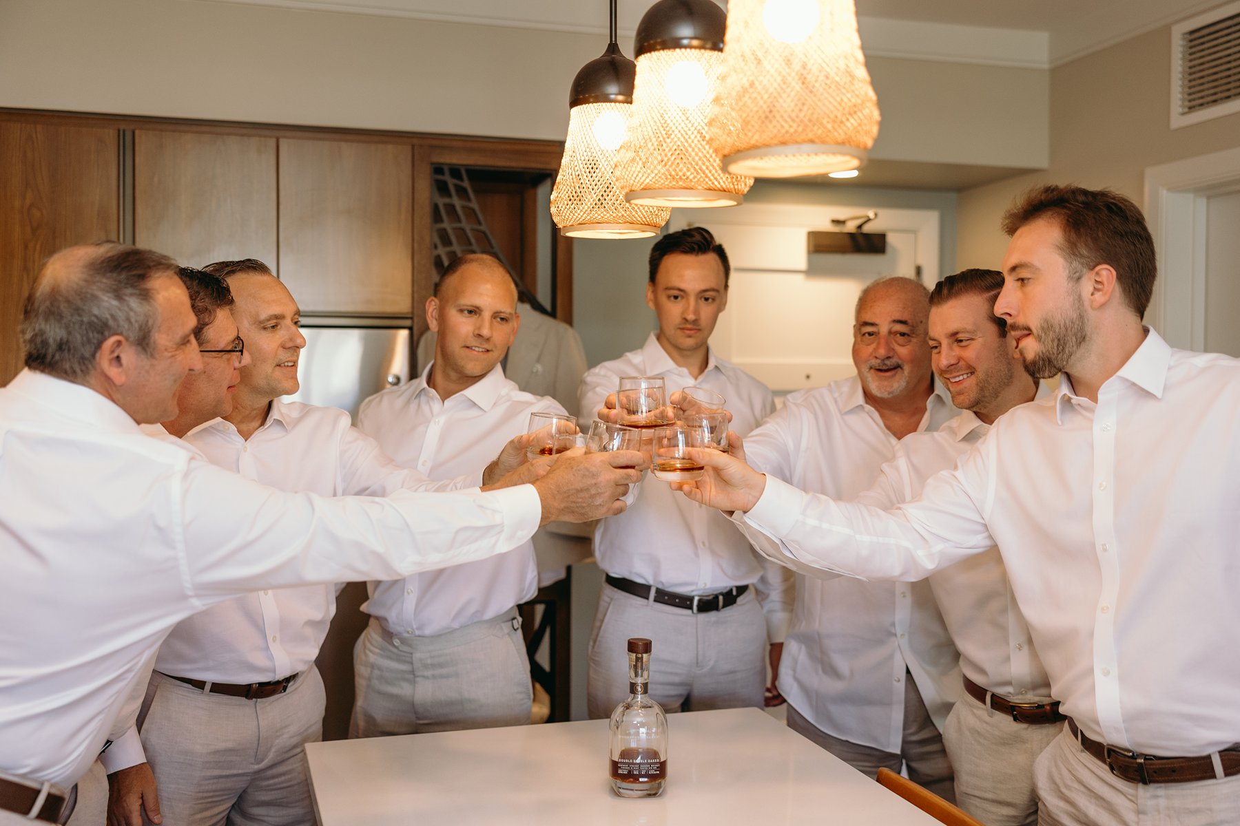Groomsmen raising a toast with whiskey glasses in a warm kitchen before their friend prepares to elope in Maui.
