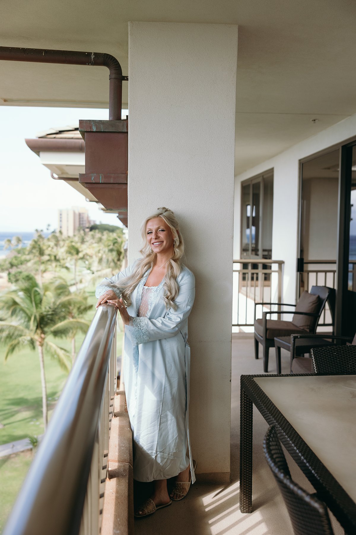 Bride standing on a resort balcony overlooking palm trees and ocean views the morning she plans to elope in Maui, soft light wrapping around her blue robe.