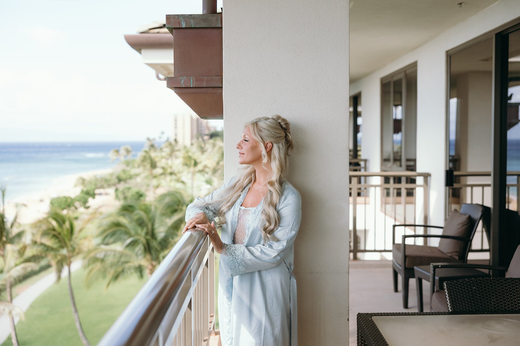 Bride standing on a resort balcony in a soft blue robe, looking out over palm trees and the ocean on the morning she will elope in Maui.