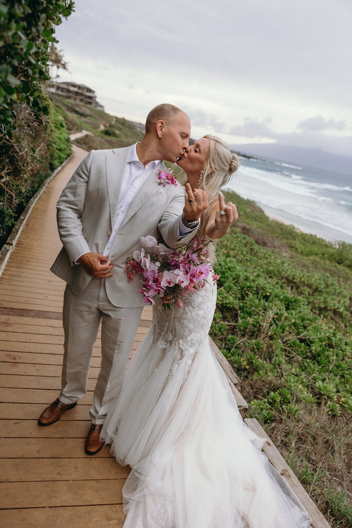 Bride and groom kissing on a coastal boardwalk after they elope in Maui, ocean cliffs and rolling waves stretching behind them as she shows off her ring and pink bouquet.