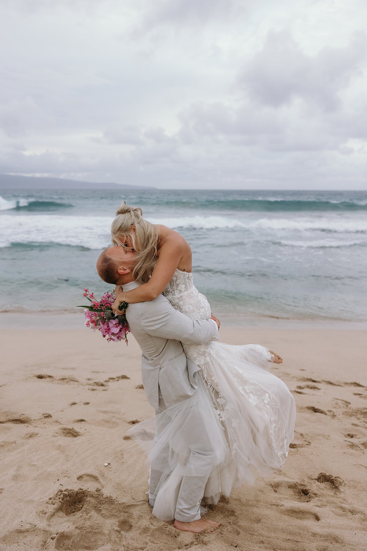 Romantic beach dip and kiss at sunset, bride’s veil and dress flowing in the Maui trade winds.