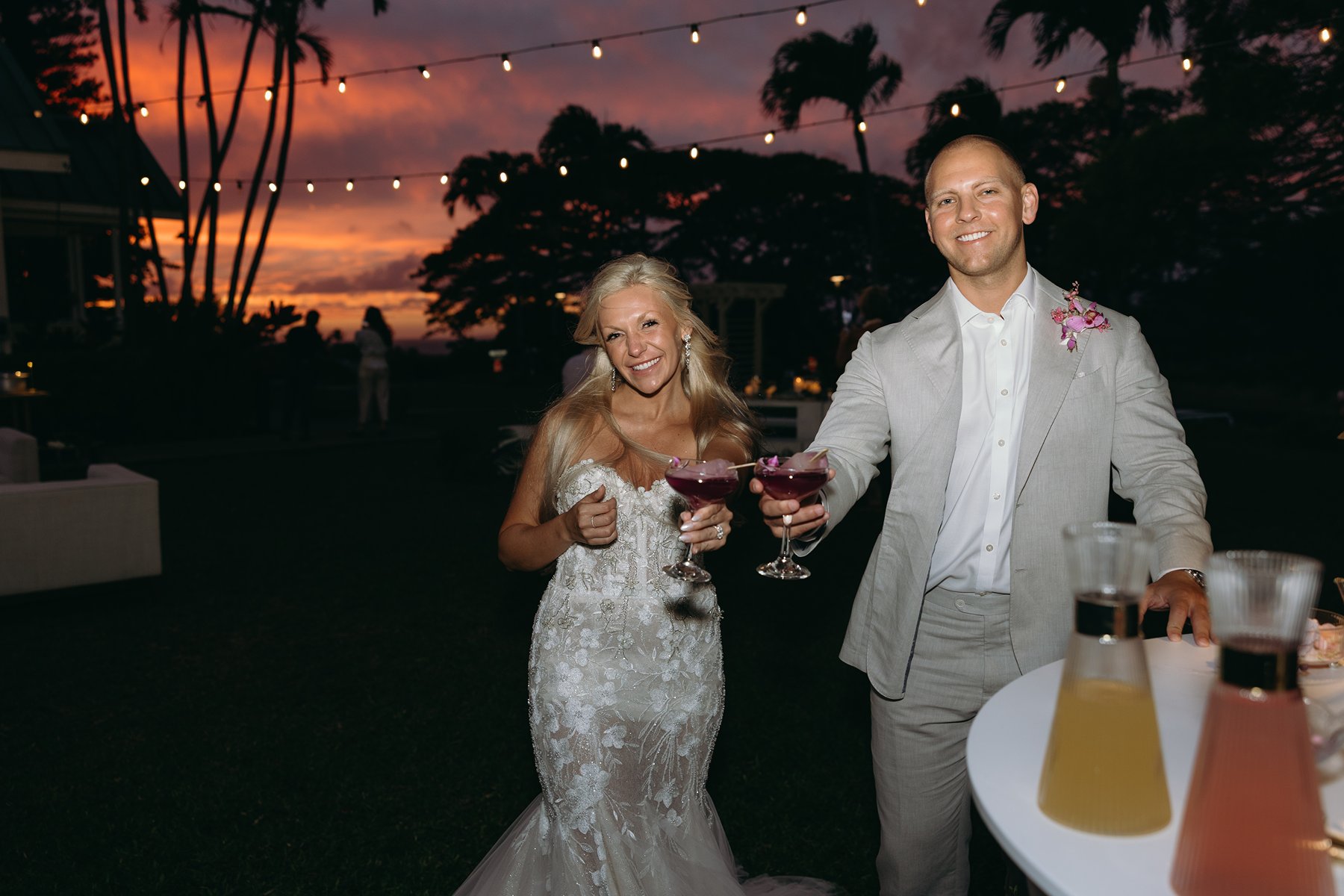 Newlyweds holding purple cocktails under glowing string lights at sunset, celebrating their Maui wedding as palm trees silhouette against a fiery sky.