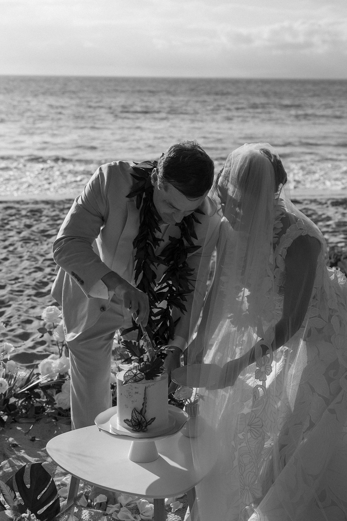 Black and white photo of bride and groom cutting their wedding cake together at a beach ceremony setup.