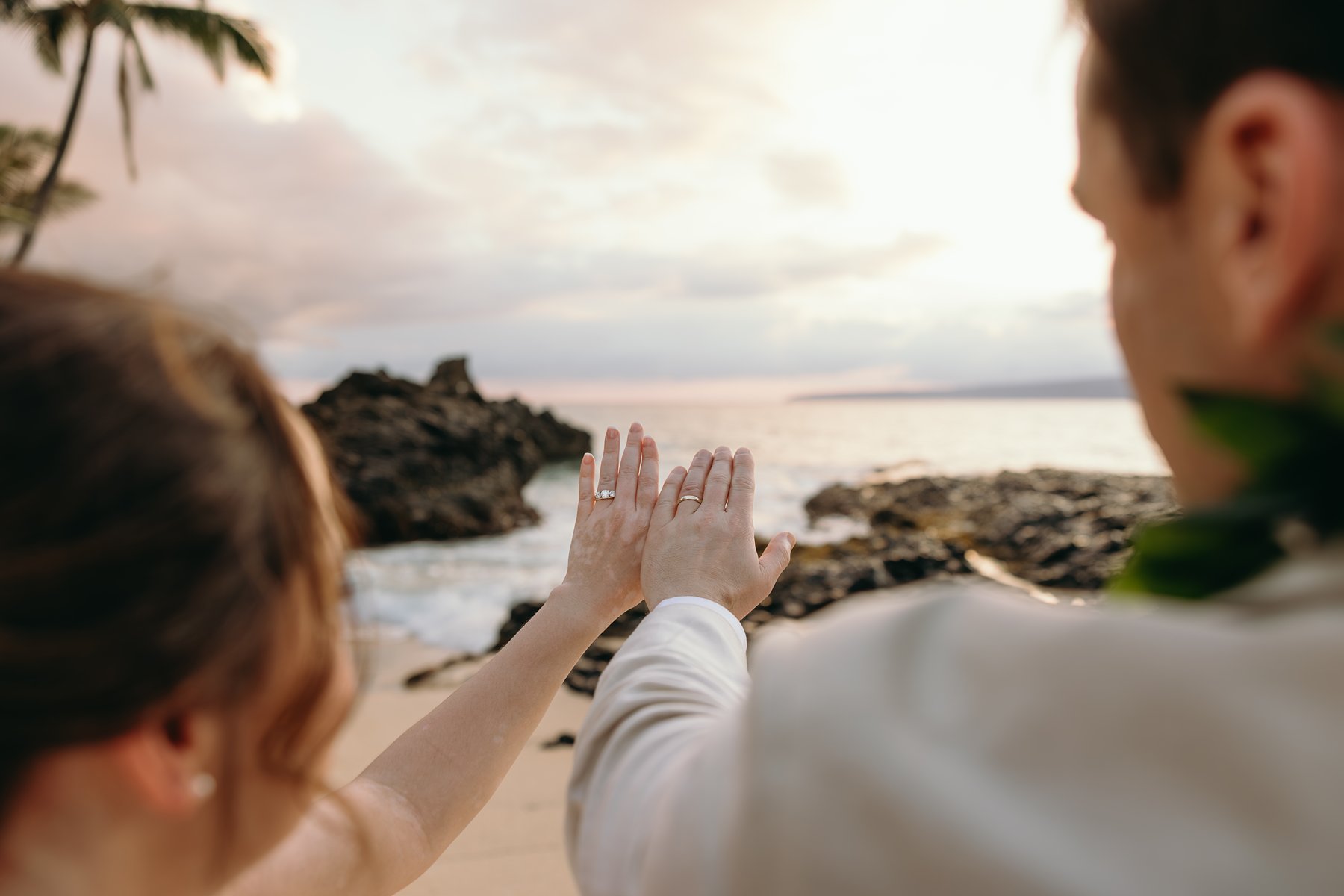 Close-up of bride and groom showing their wedding rings with ocean waves and volcanic rocks in the background.