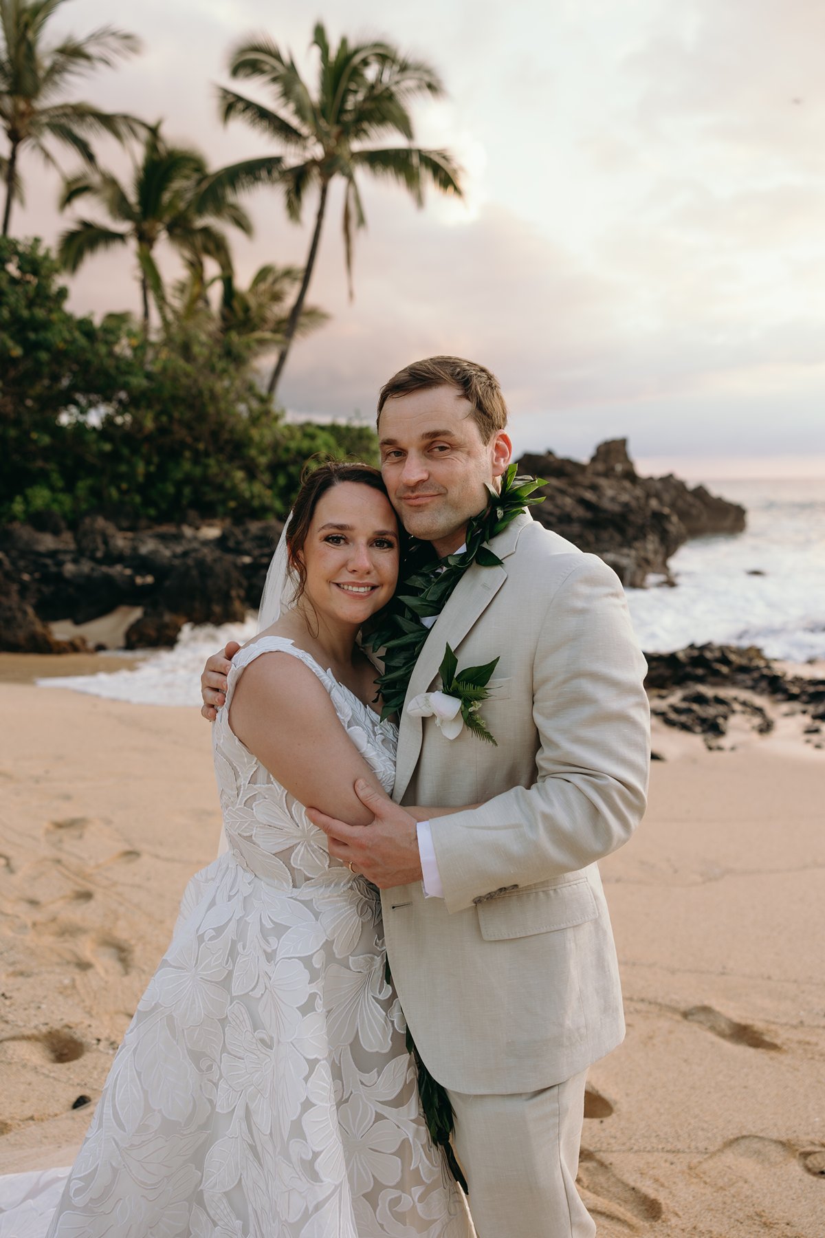 Bride and groom embracing on a golden sand beach with palm trees and ocean waves behind them.