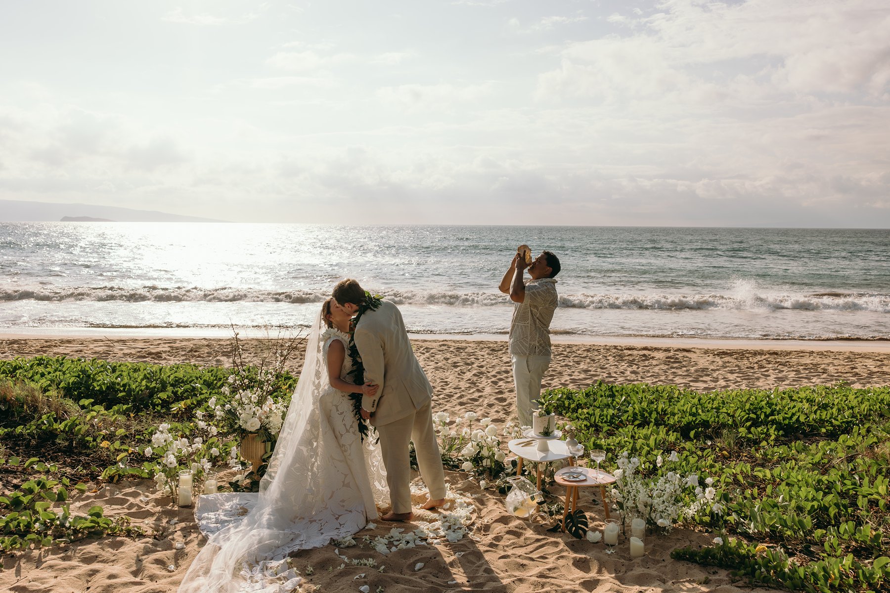 Hawaiian officiant blowing a conch shell as the couple kisses during their Maui beach ceremony captured in elopement photography.