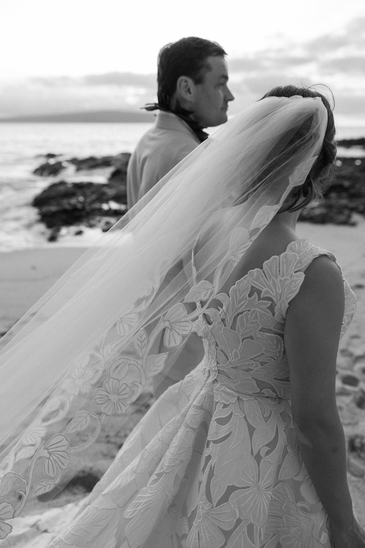 Black and white portrait of a bride with a flowing veil standing beside her groom on a Maui beach.