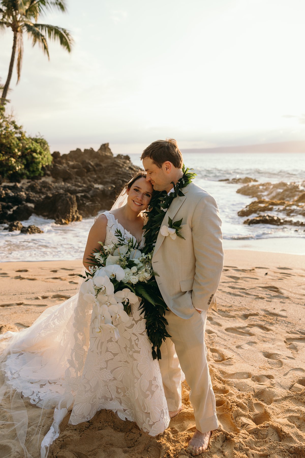 Bride and groom embracing barefoot on a sandy Maui beach with palm trees and lava rocks in the background.