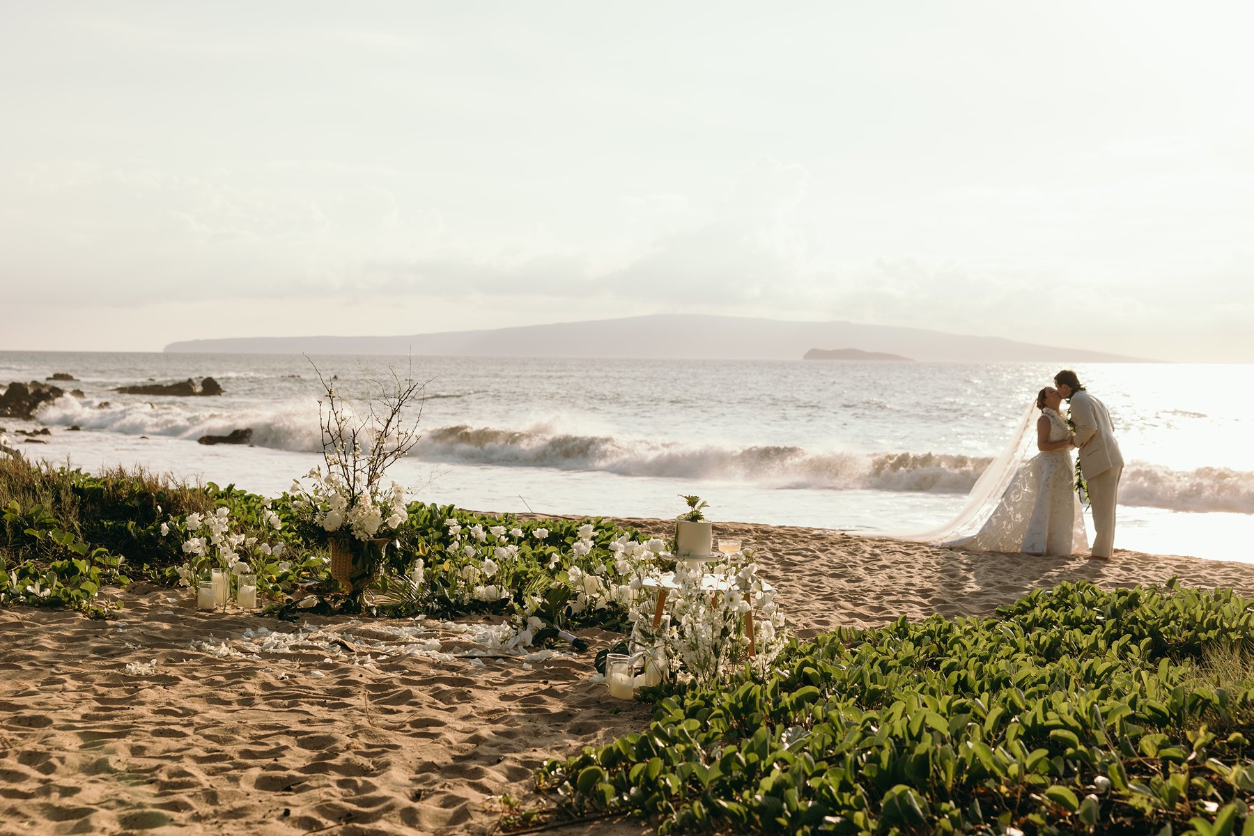 Romantic Maui beach ceremony setup with white florals and candles as the couple kisses by the ocean during elopement photography.