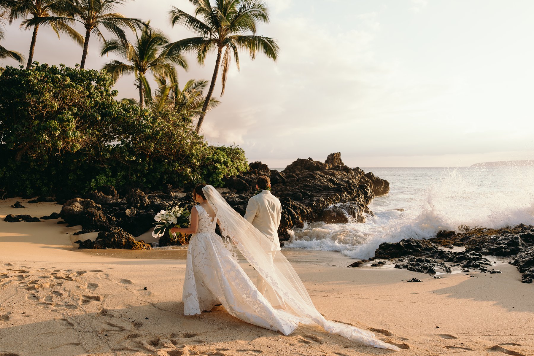Bride and groom walking along a secluded Maui beach with palm trees and crashing waves during golden hour elopement photography.