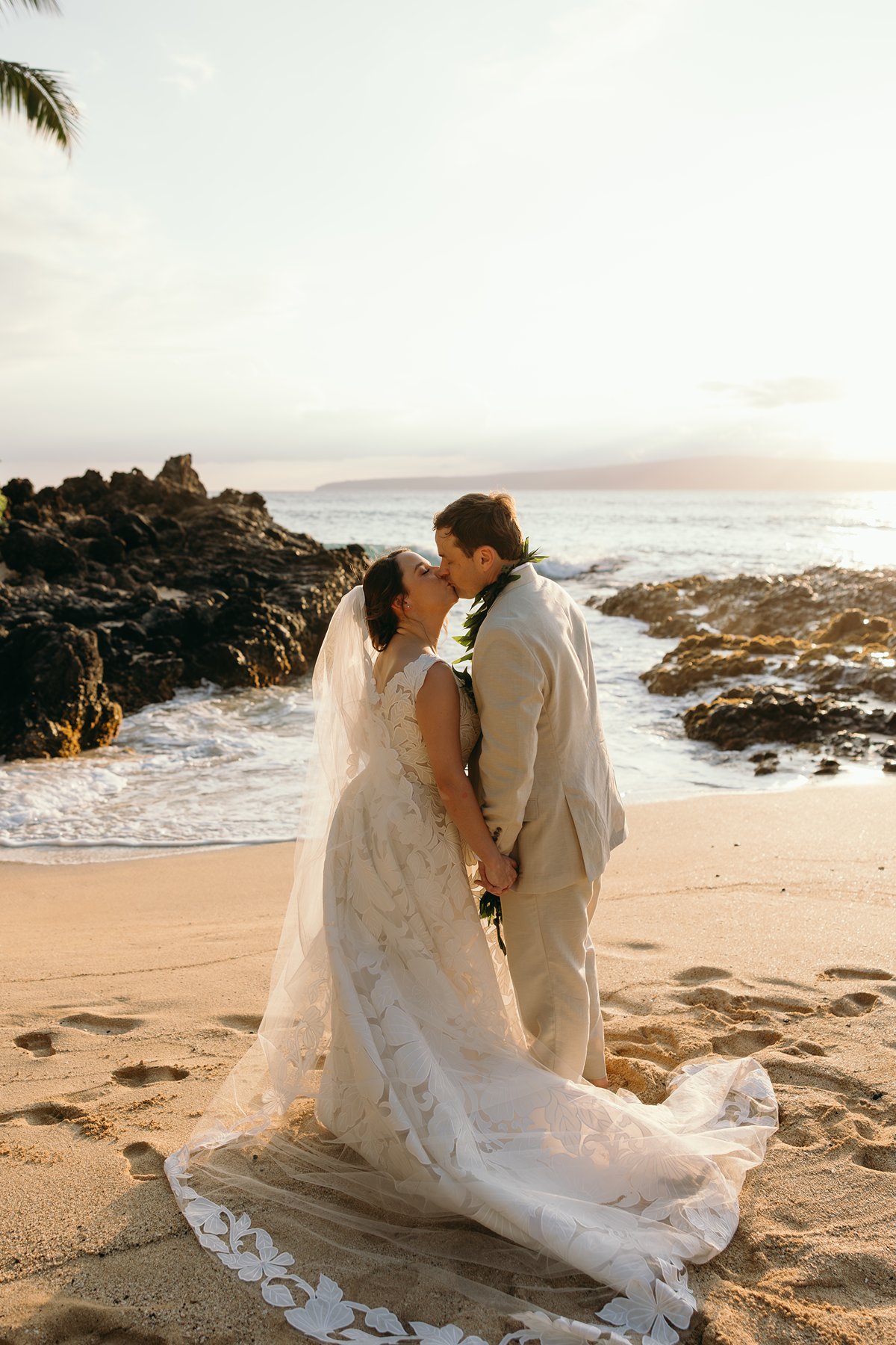 Bride and groom sharing a kiss on a golden sand beach with lava rocks and ocean waves in the background.