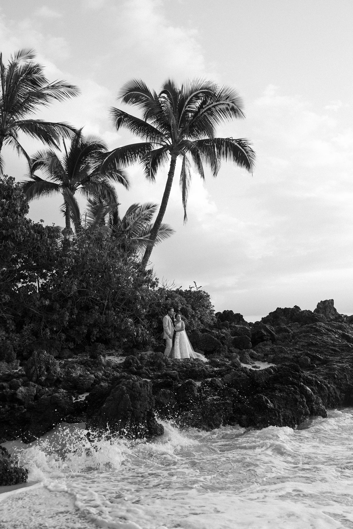Black and white photo of a couple standing on volcanic rocks beneath tall palm trees along the Maui coastline during elopement photography.