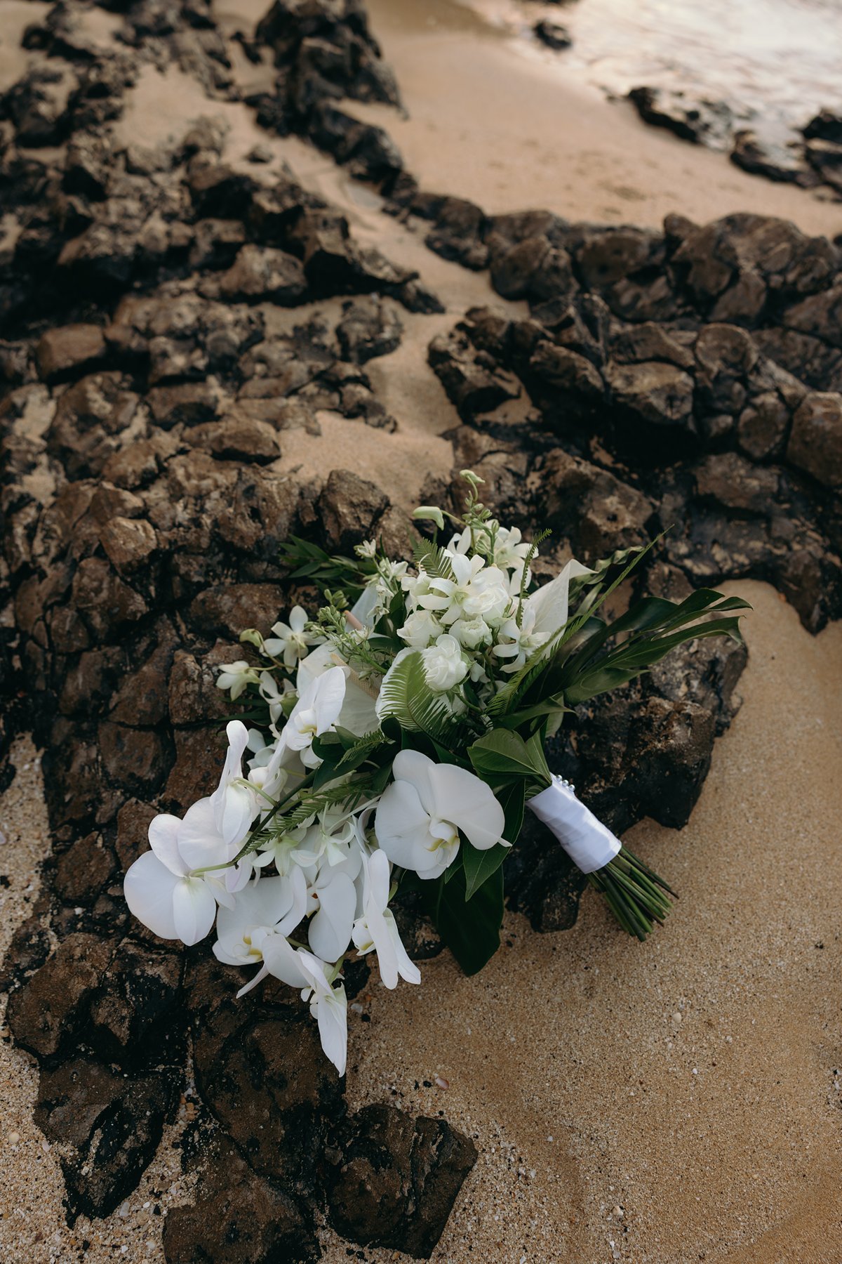 White tropical orchid wedding bouquet resting on volcanic lava rocks along a Maui shoreline.