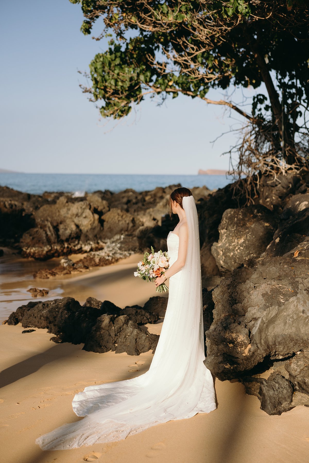 Bride standing by ocean rocks at sunset in glowing wedding sneak peek photos